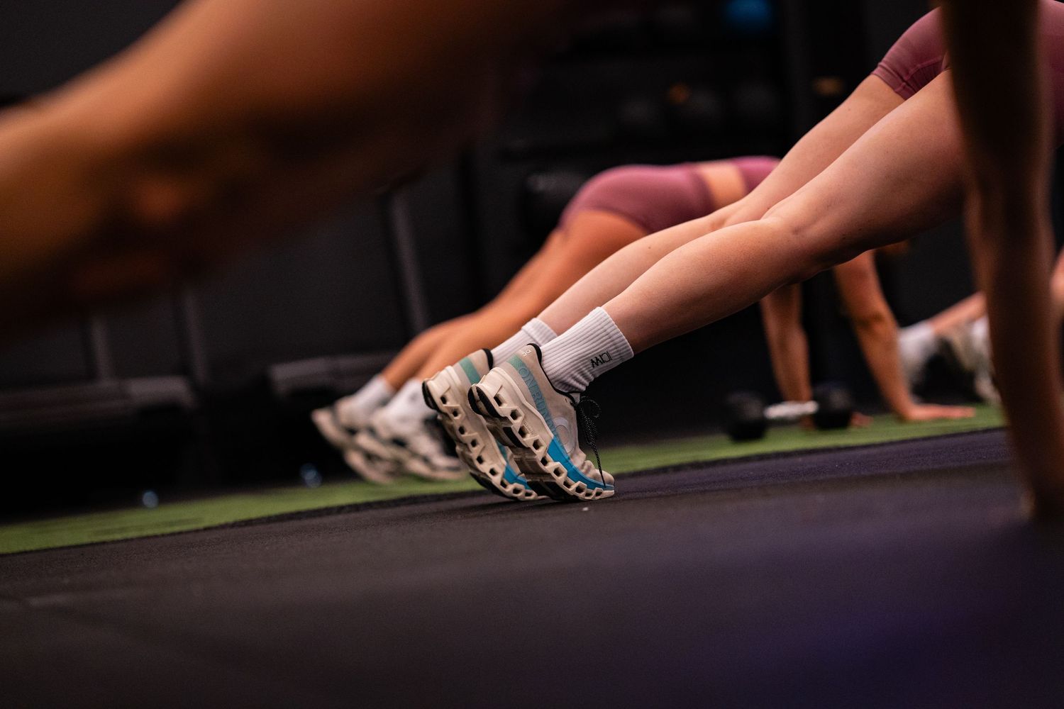 People performing push-ups in a gym, focusing on athletic shoes and legs on a dark exercise floor.