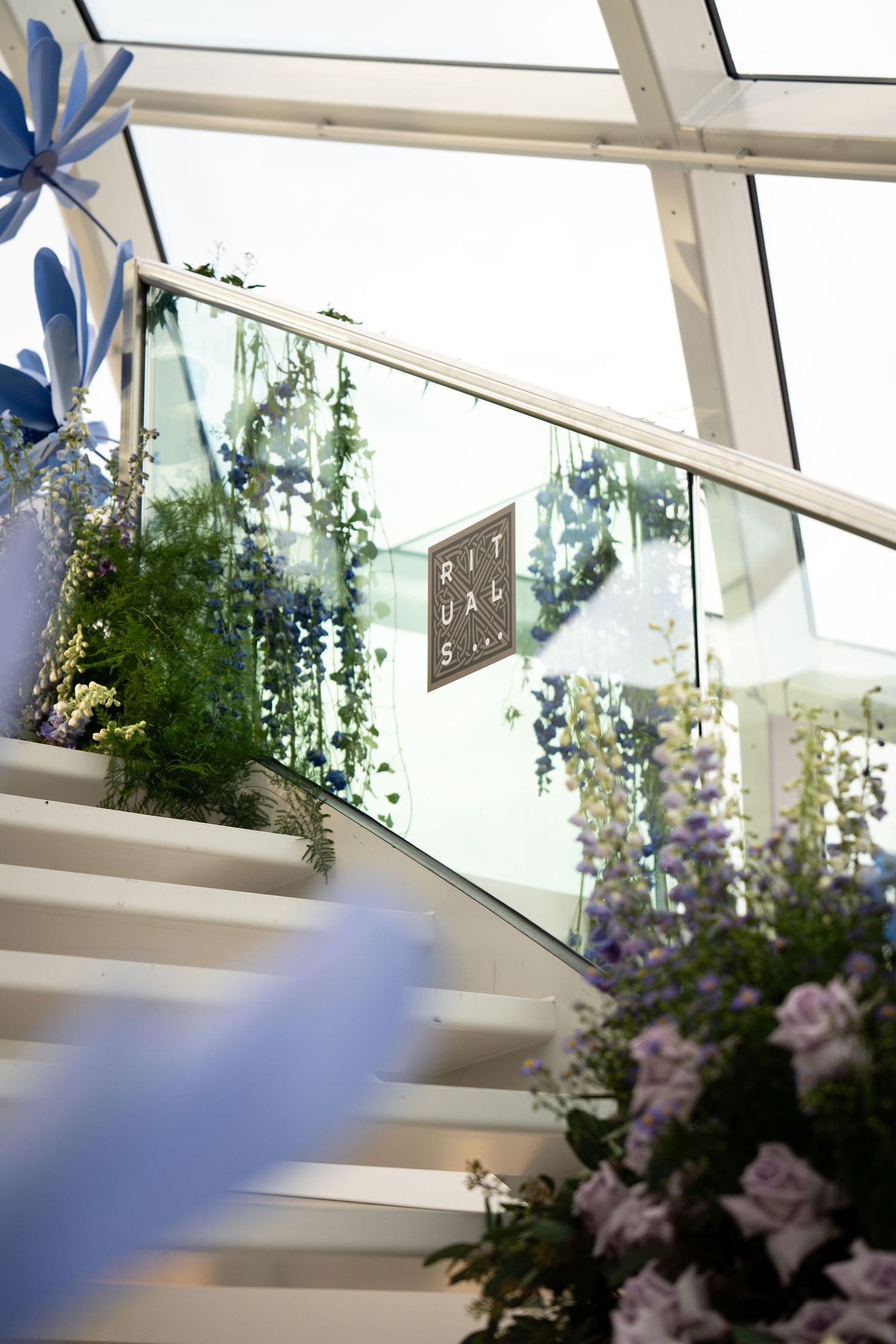 Rituals store staircase adorned with lush green plants and purple flowers, featuring glass railings in a bright modern space.