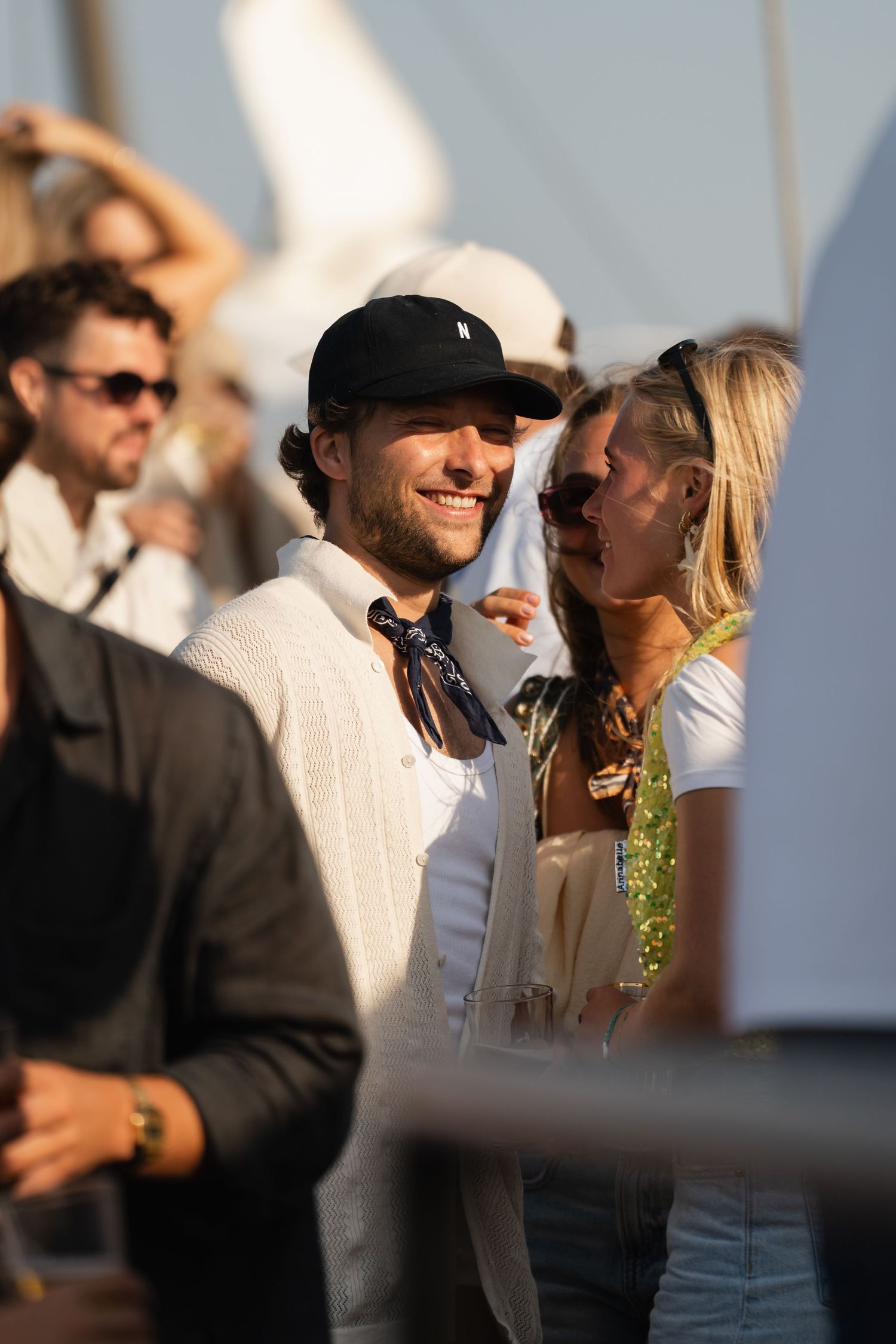 Smiling man in black cap and white jacket with bandana at an outdoor social event.