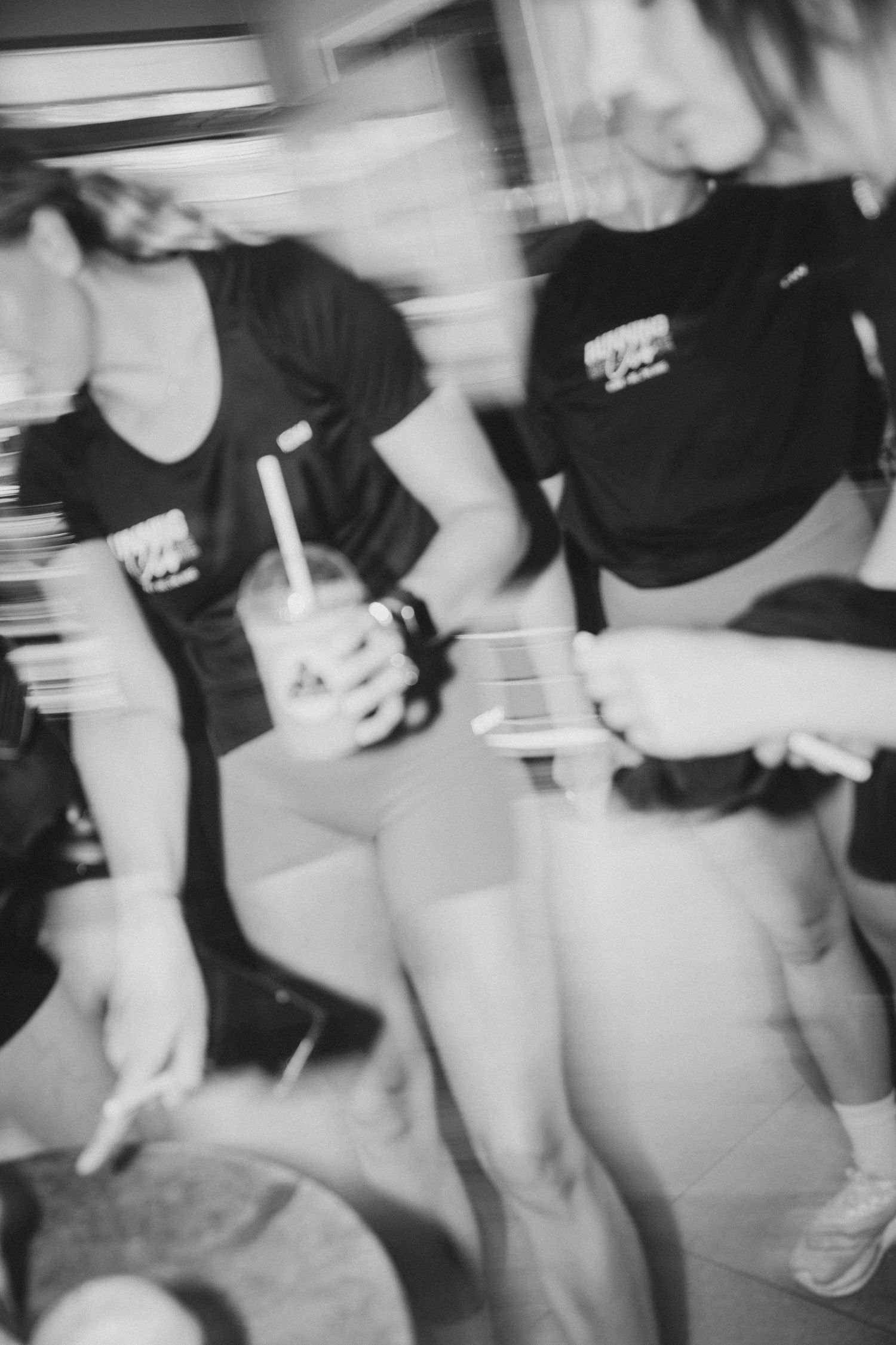 Blurry black and white photo of two women in matching black t-shirts, one holding a drink with a straw.