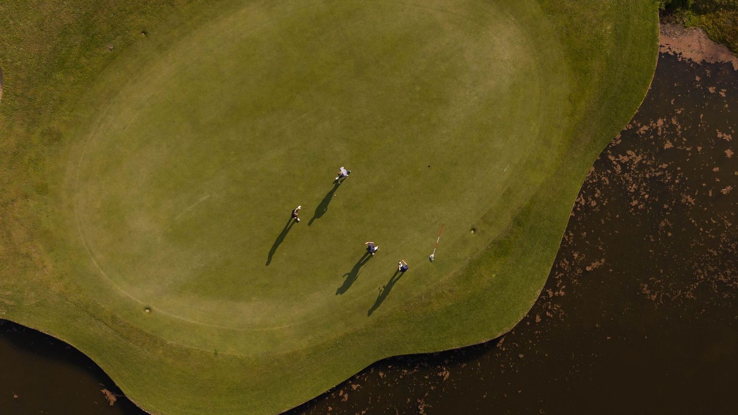 Aerial view of three golfers on a lush green golf course beside a water hazard, casting long shadows.
