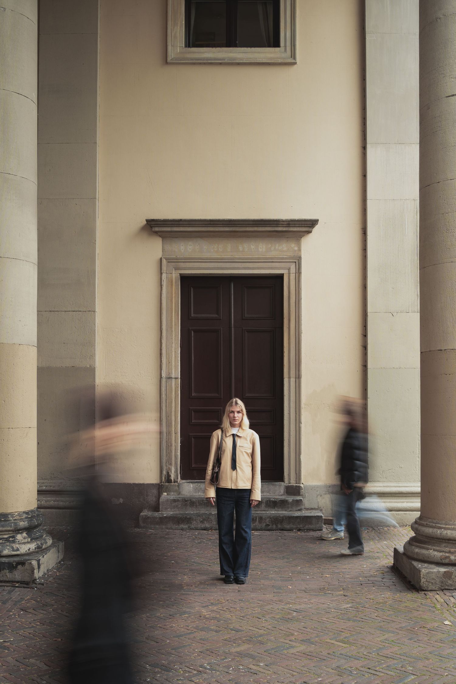 Woman in beige jacket stands still before ornate door as blurred figures pass by classical building columns.