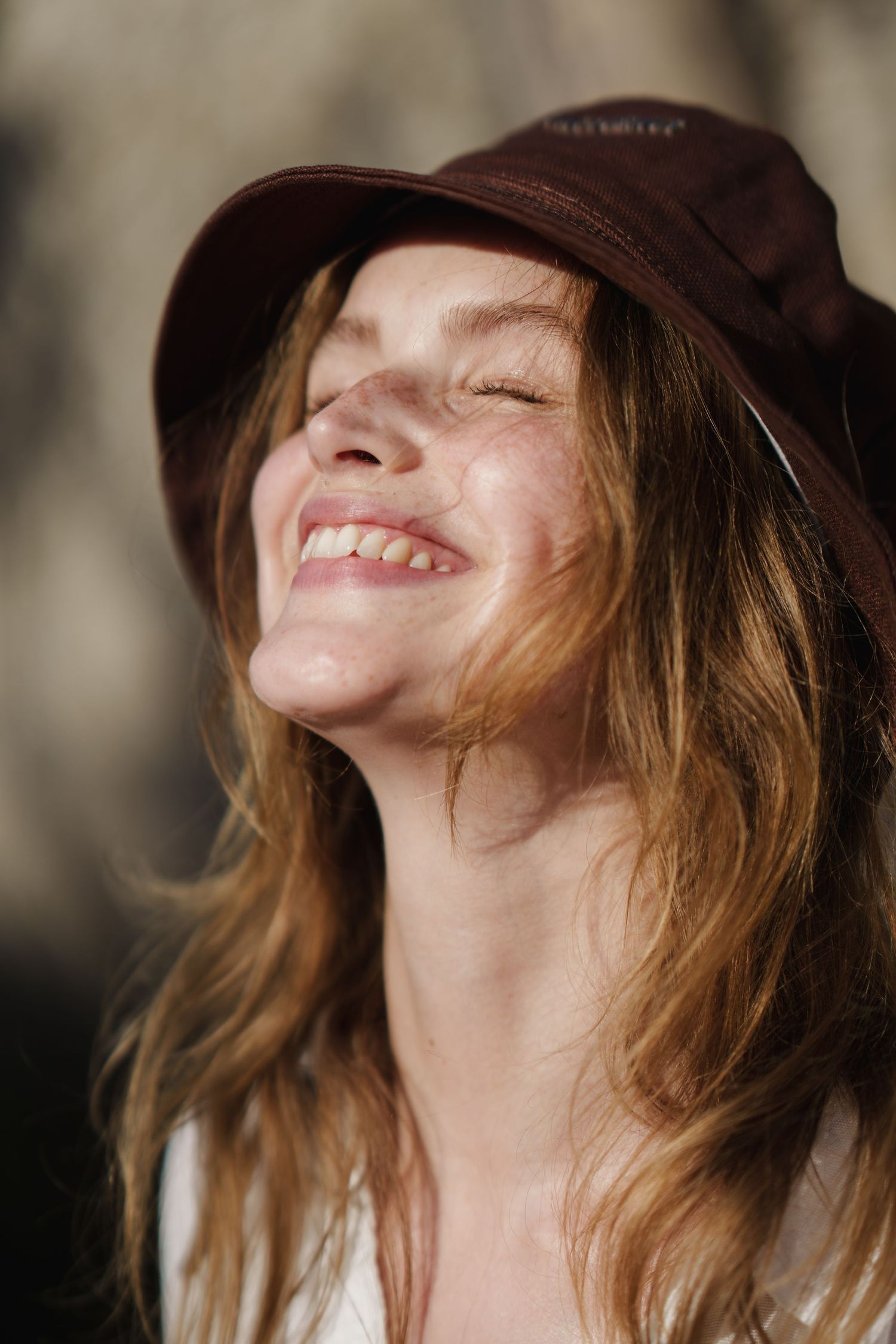 Smiling woman with freckles and long auburn hair wearing a dark burgundy bucket hat in natural sunlight.