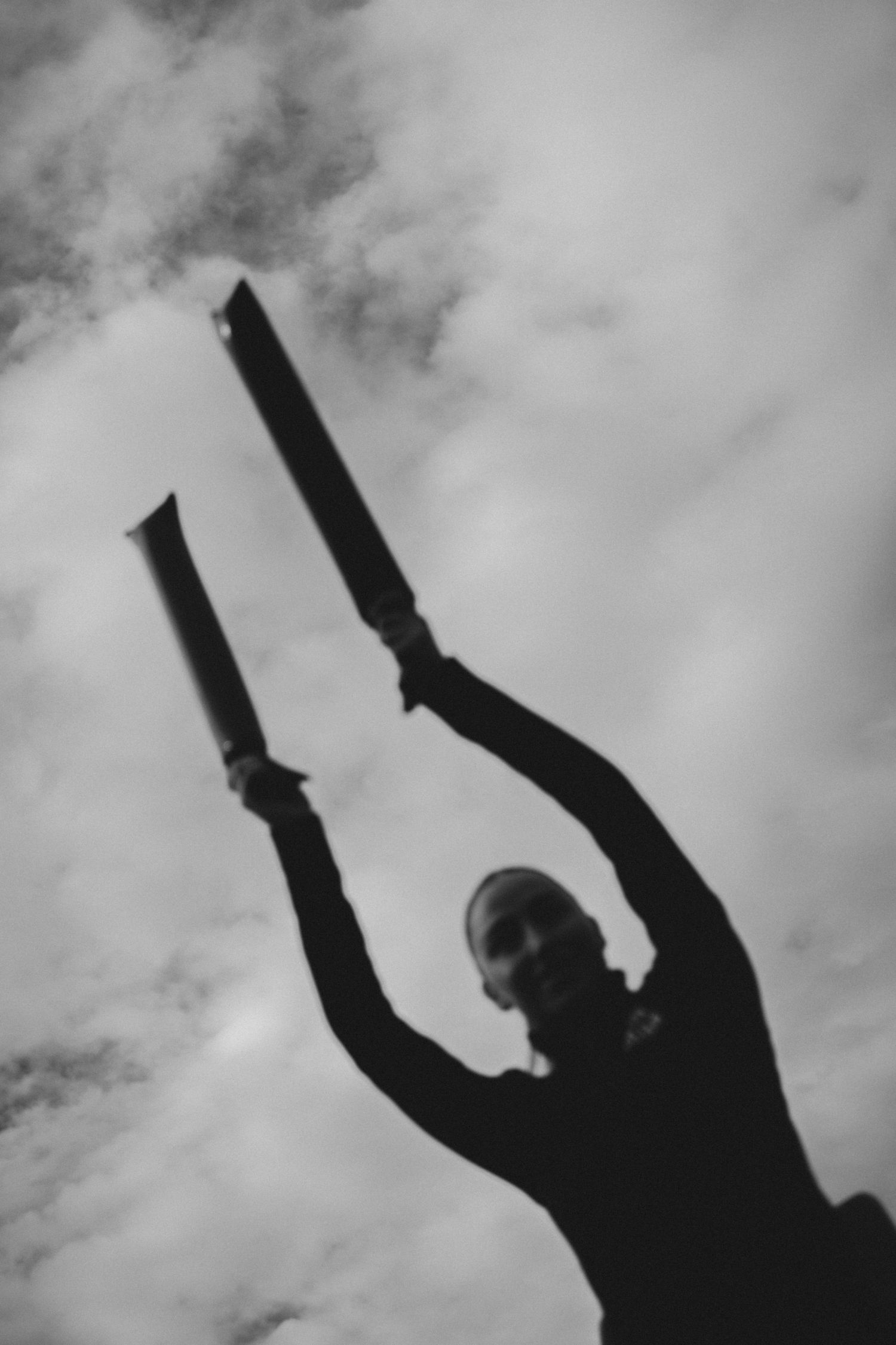 Silhouette of person holding batons against cloudy sky in dramatic black and white.
