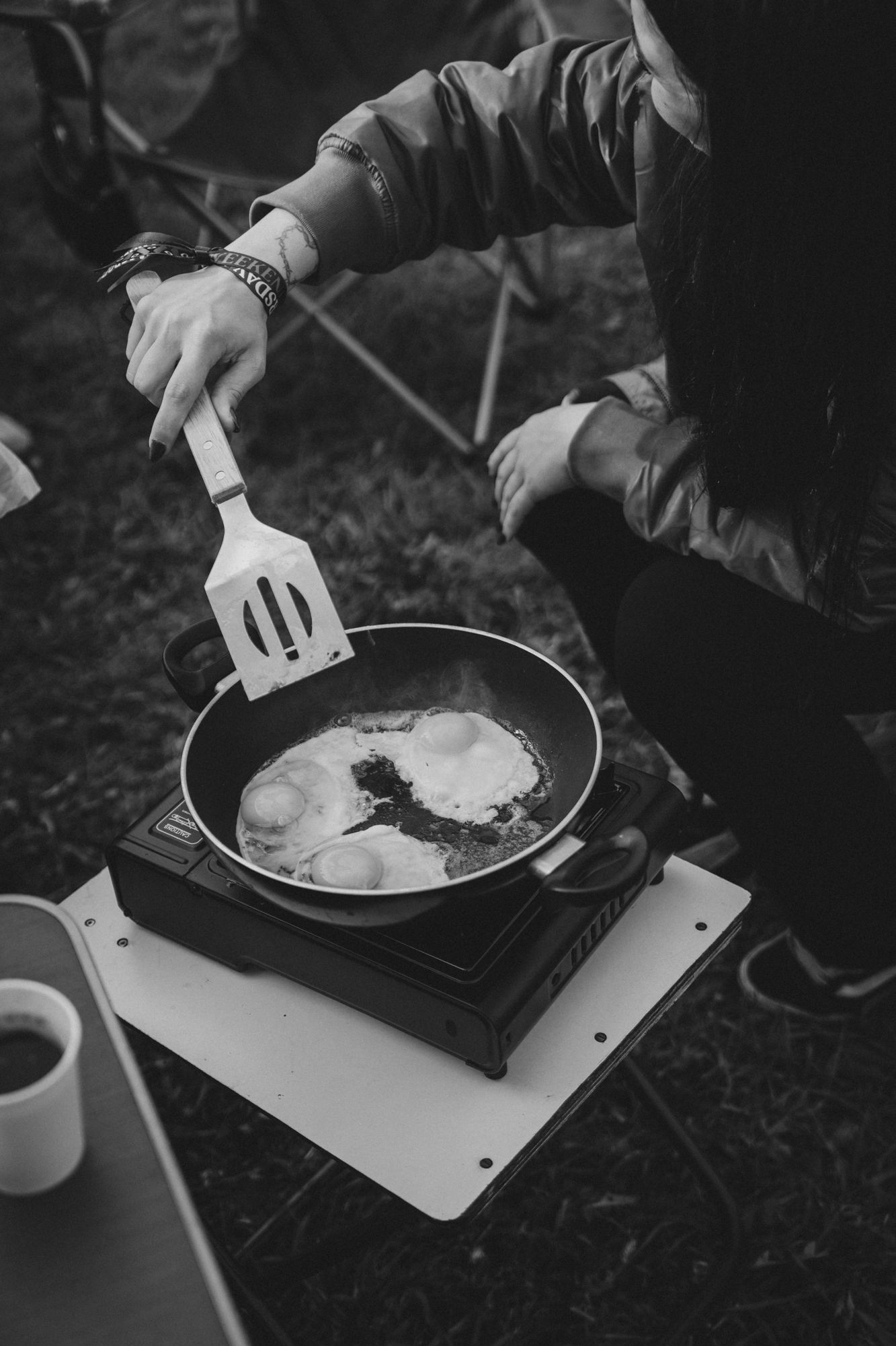 Person cooking fried eggs in a pan on a portable camping stove outdoors, black and white photography.