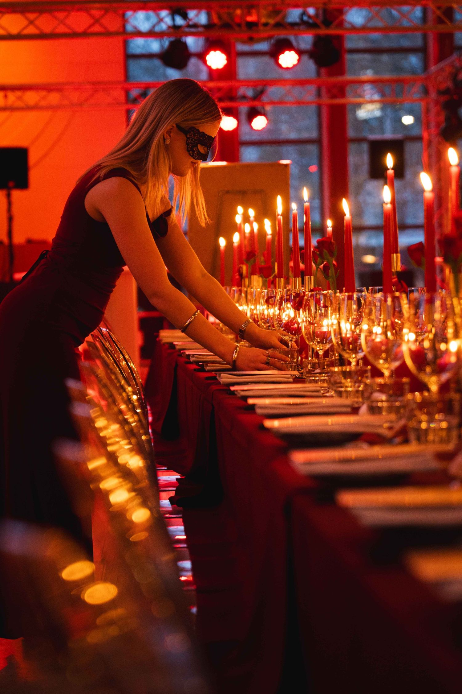 Blonde woman in black dress arranging a luxurious red-lit candlelit banquet table with wine glasses.
