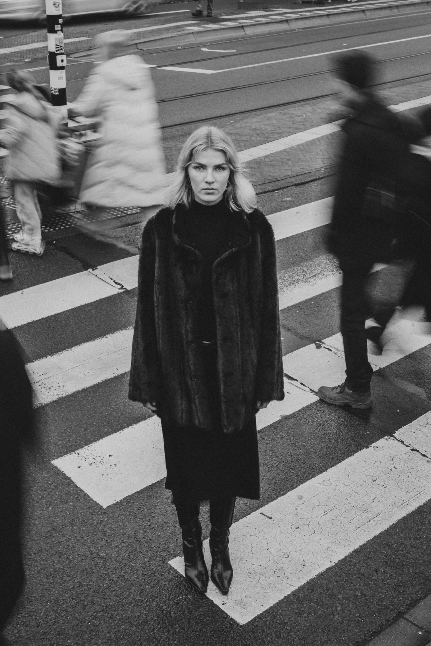 Black and white photo of a blonde woman in a fur coat standing still on a busy zebra crossing.