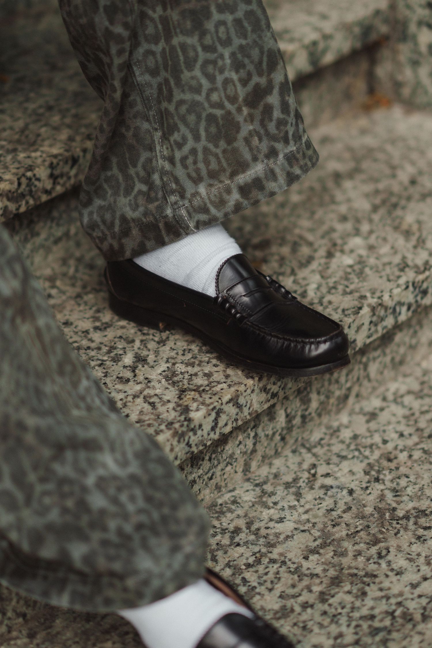 Black penny loafers with white socks paired with leopard print trousers on granite steps.