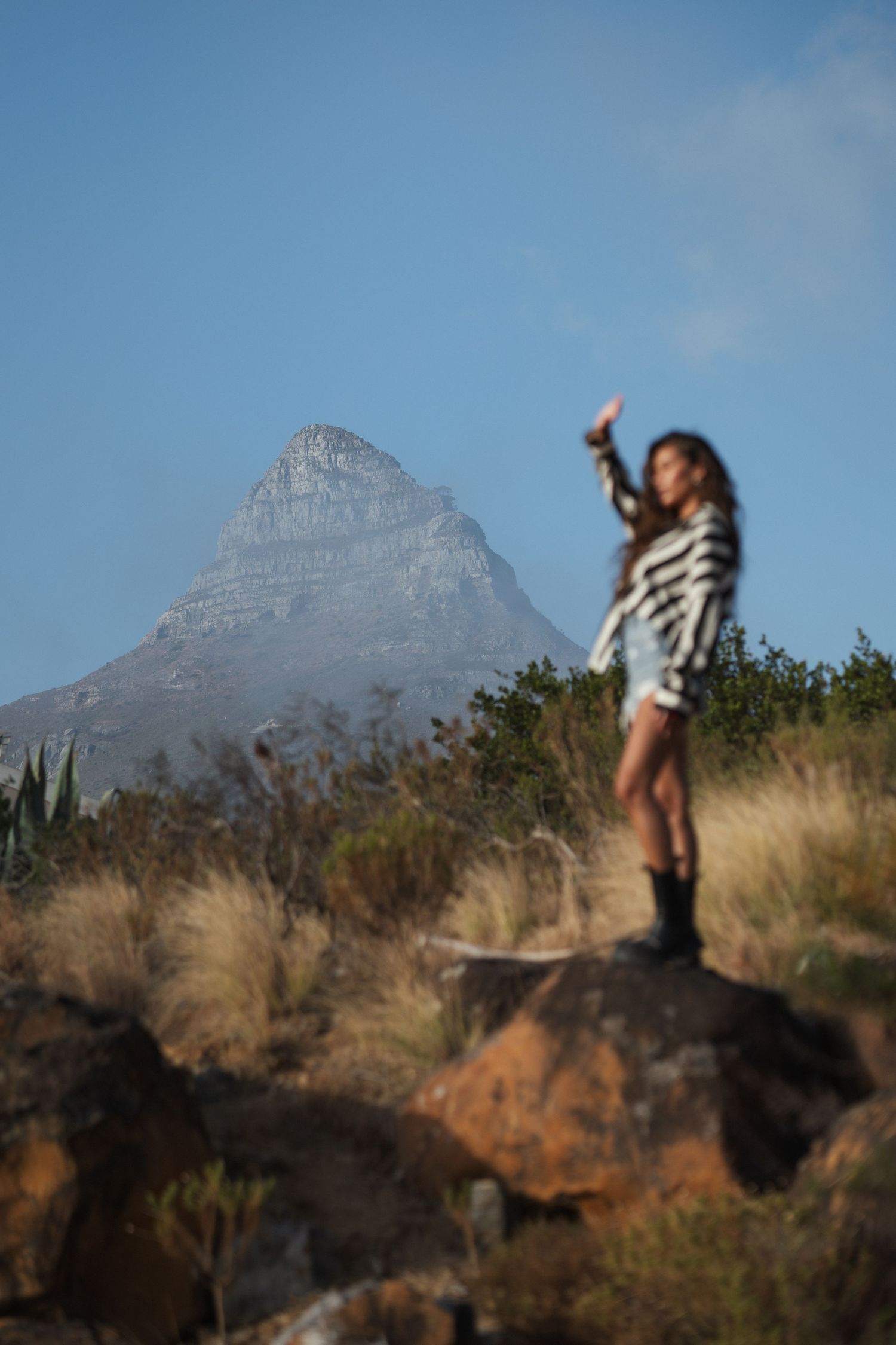 Woman standing on rocks in striped outfit with arm raised, Lion's Head mountain peak visible in background, Cape Town.