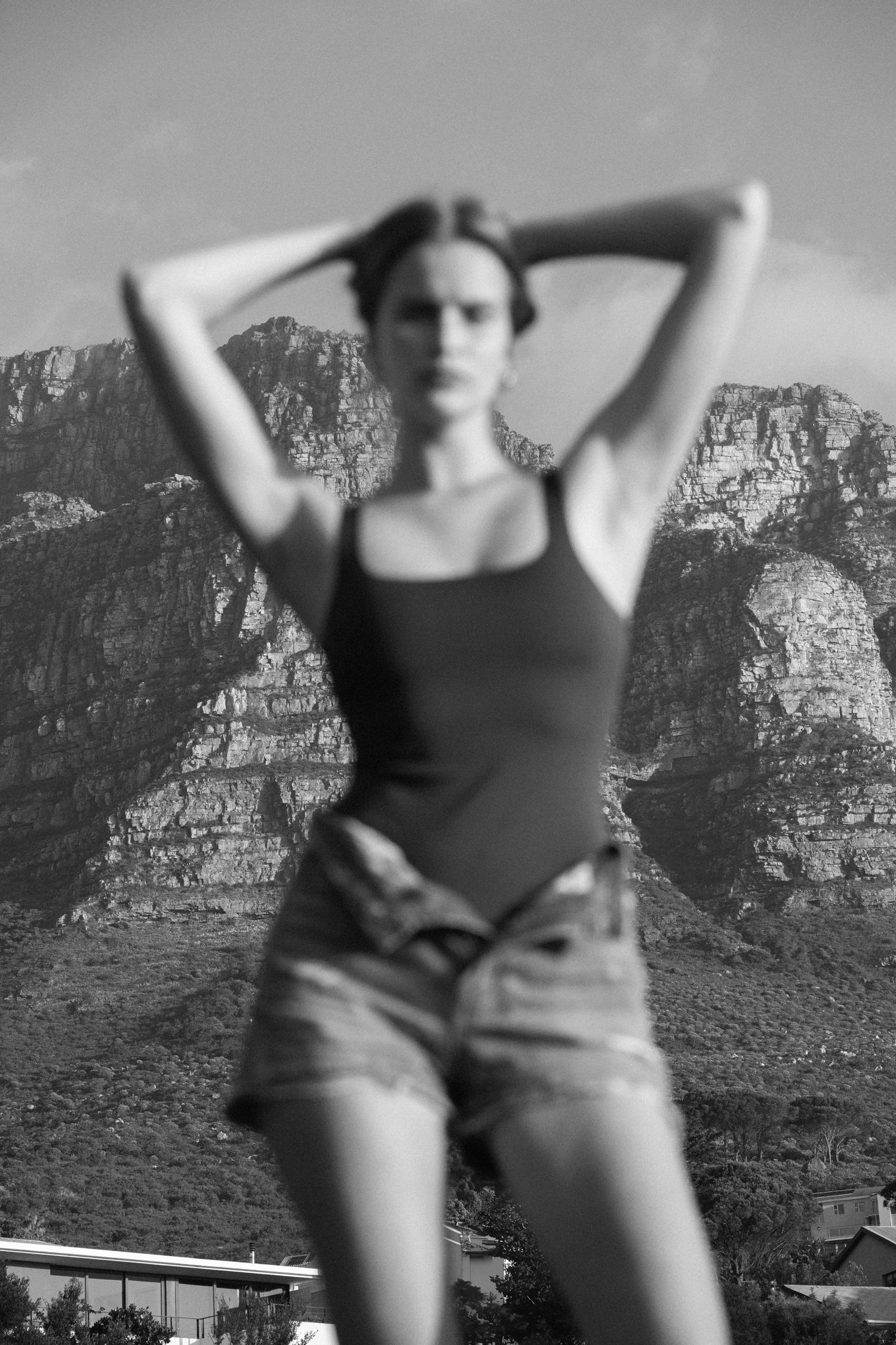 Black and white photo of a woman in tank top and denim shorts posing with hands behind head, mountains in background.