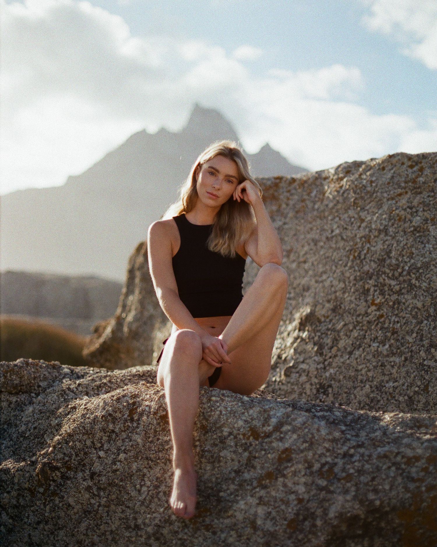 Blonde woman in black crop top sitting on rocks with dramatic mountain peaks and cloudy sky in background.
