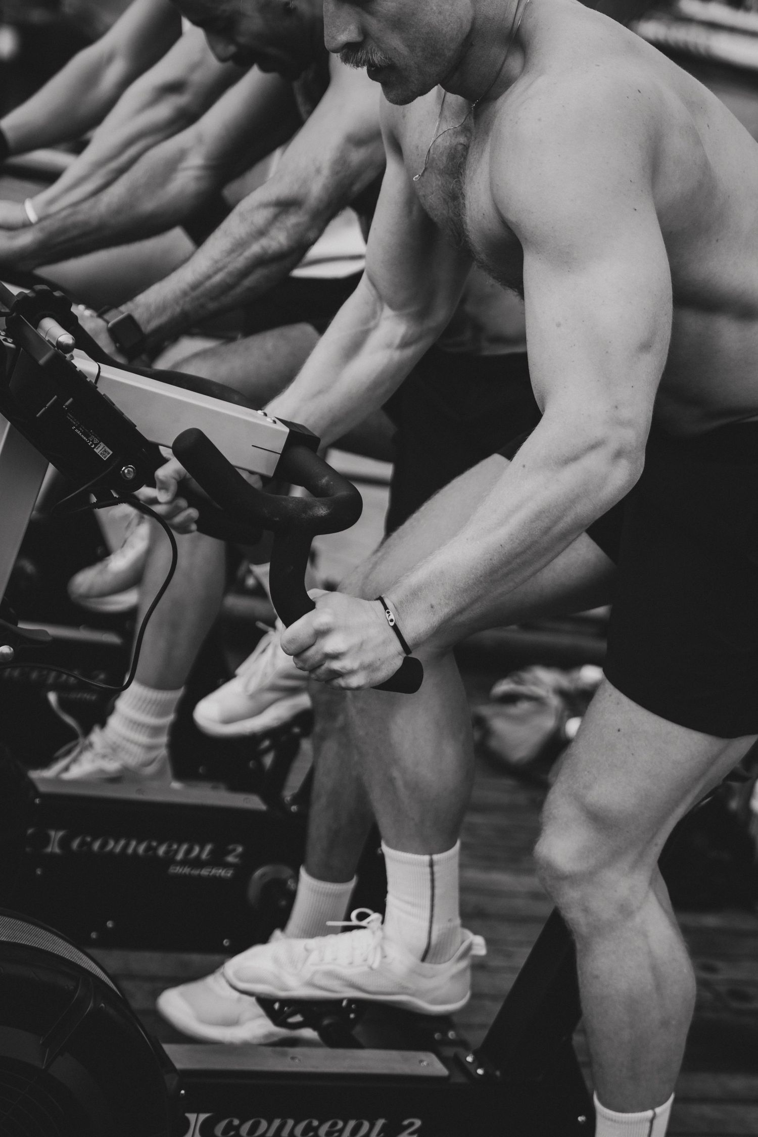 Muscular shirtless men training on Concept2 rowing machines in a gym, captured in black and white.