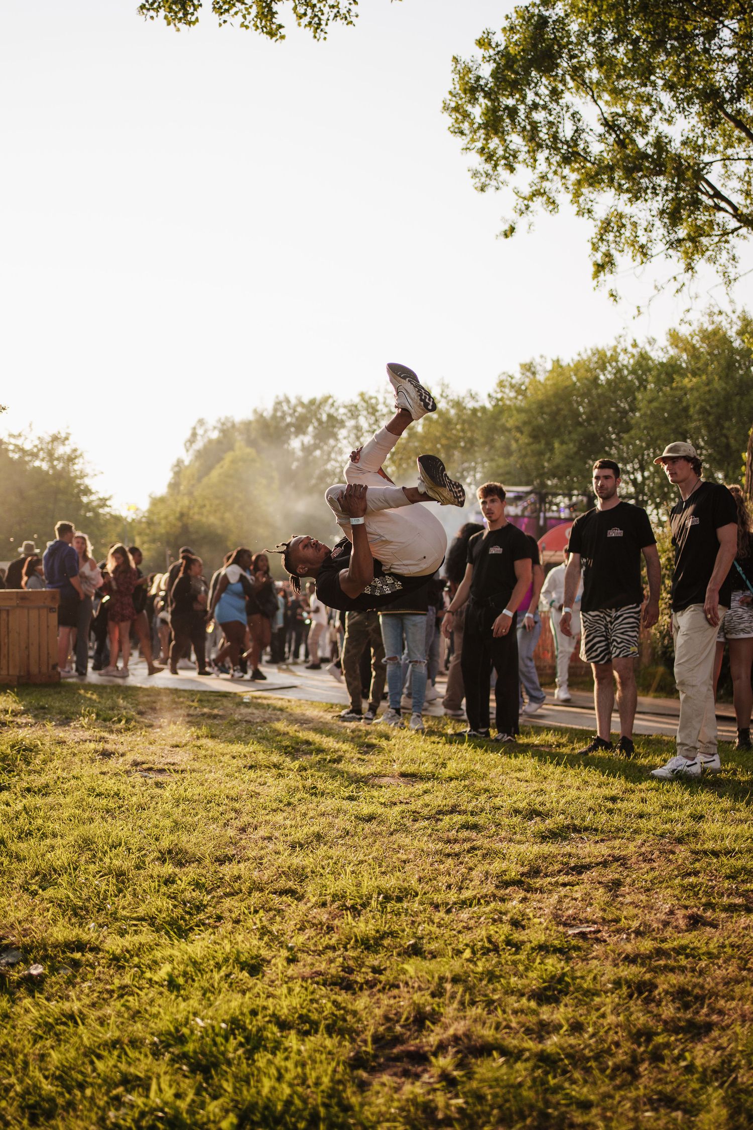 Person performing a backflip at an outdoor festival with crowds and trees in golden hour sunlight.