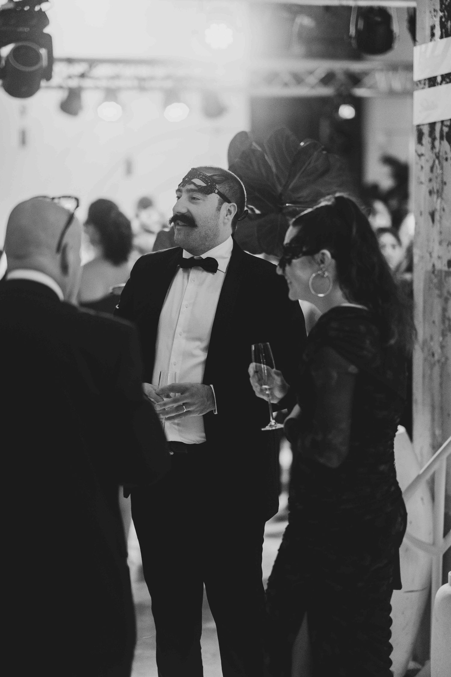Black and white photo of masked guests in formal attire socializing at an elegant masquerade ball event.