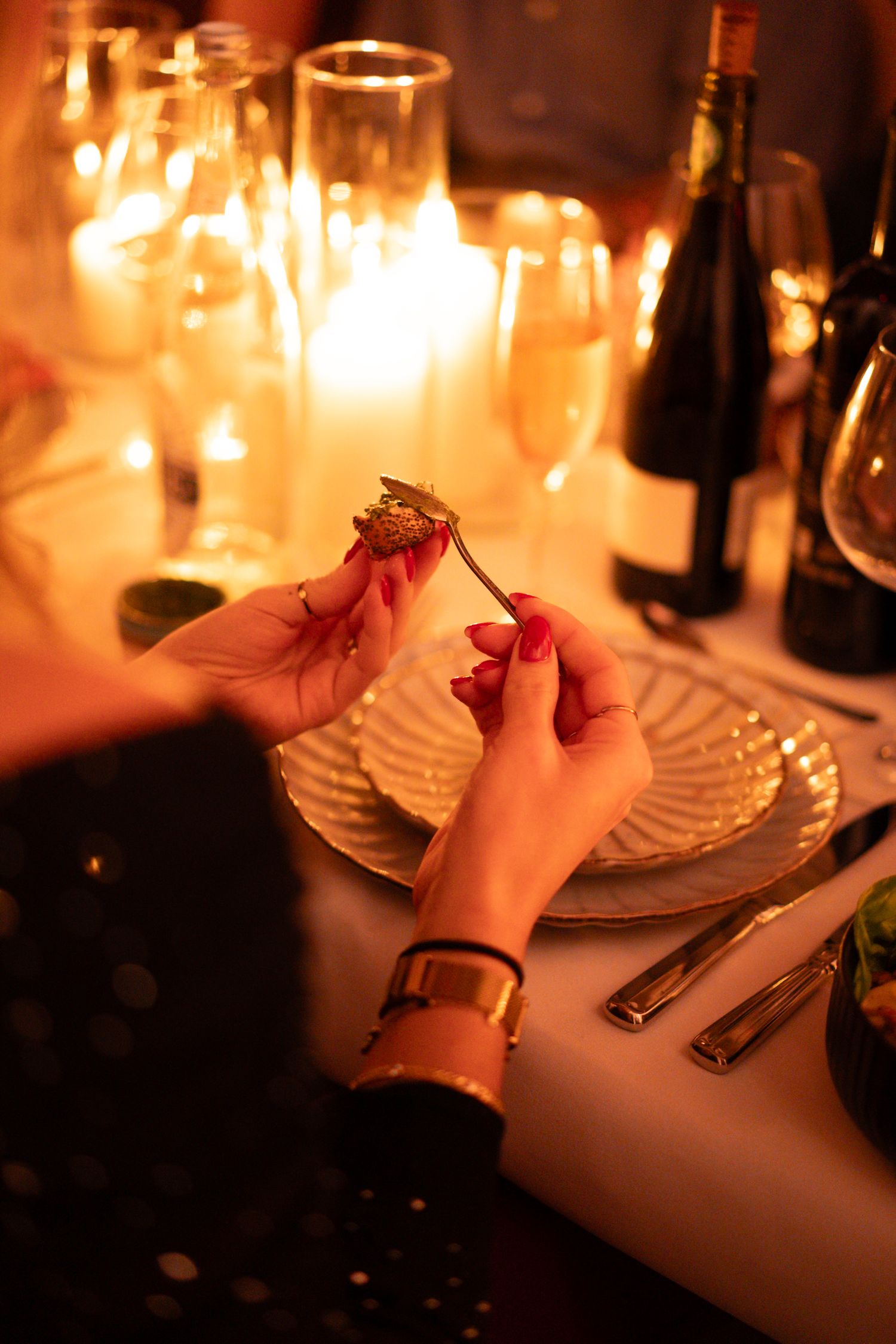 Woman with red nails holds a strawberry on a fork at a candlelit dinner table with wine glasses.