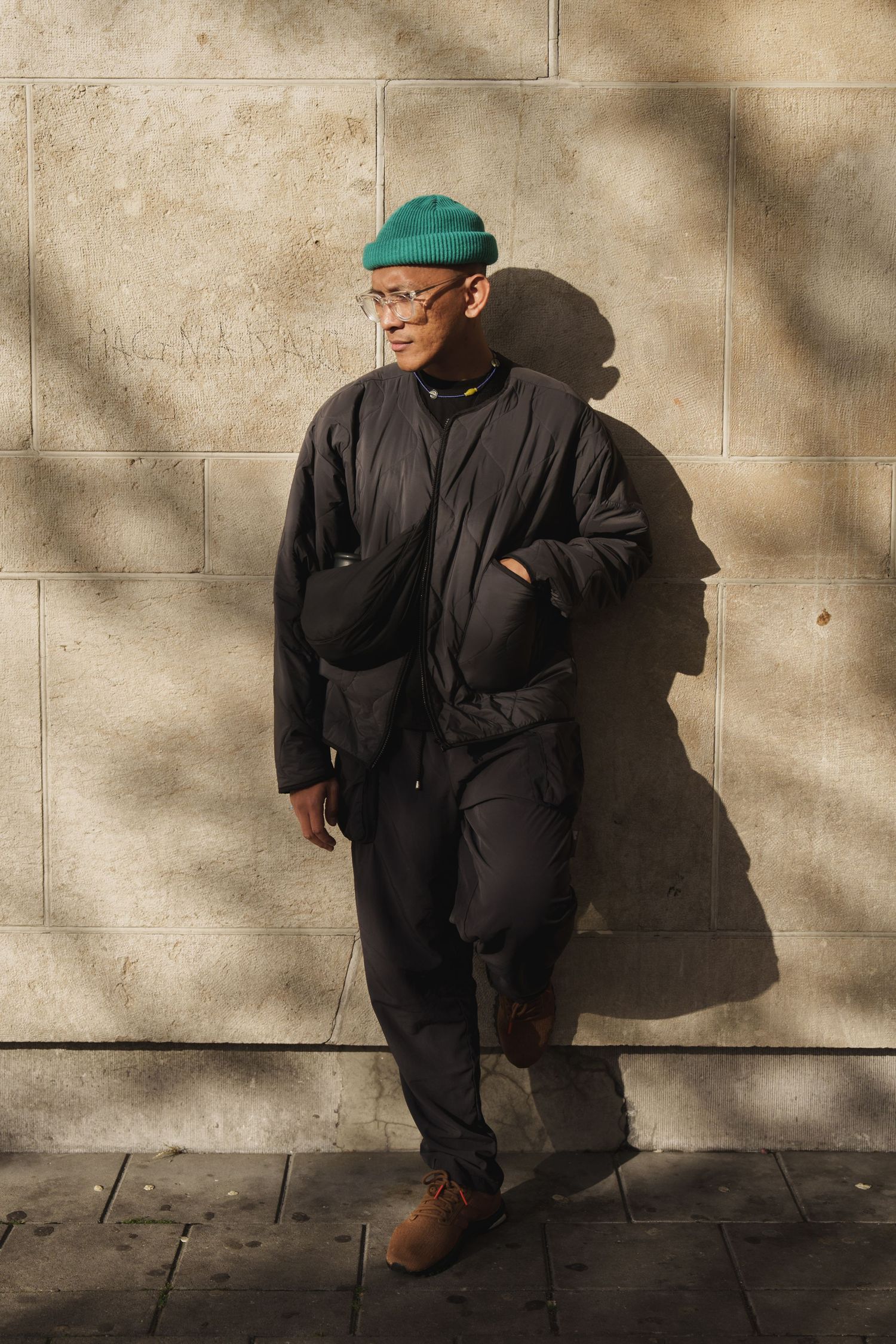 Man in black outfit and green beanie hat leaning against stone wall in sunlight.