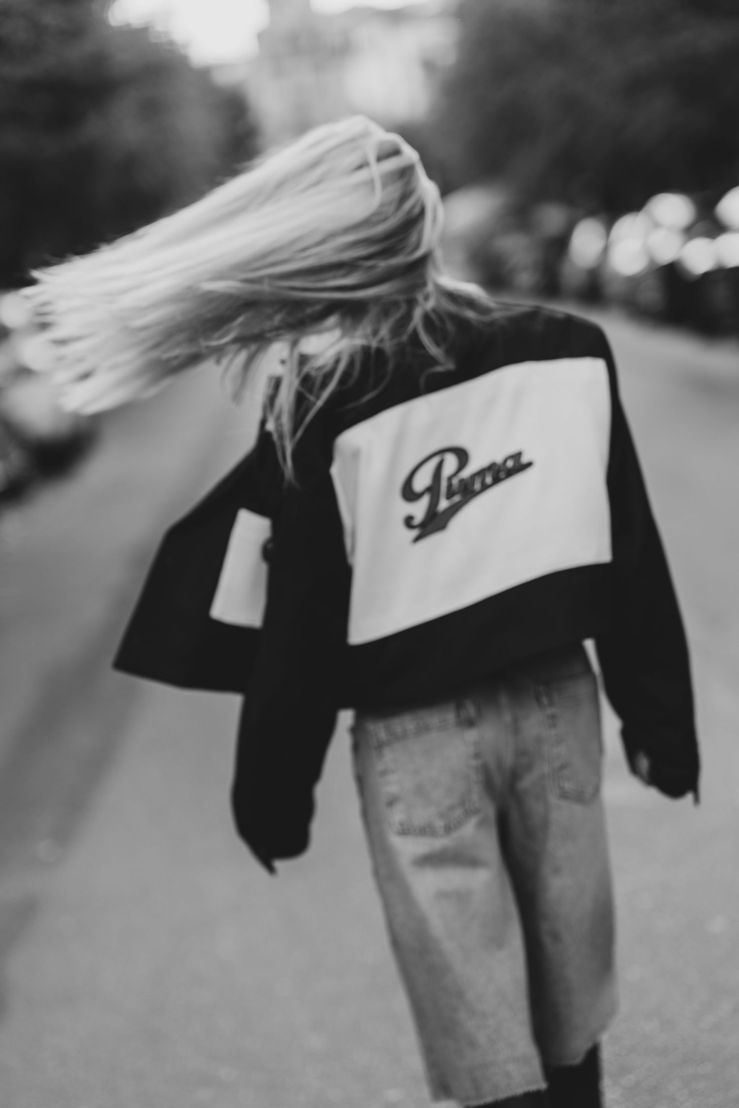 Black and white photo of a person walking away wearing a Puma varsity jacket with flowing blonde hair.