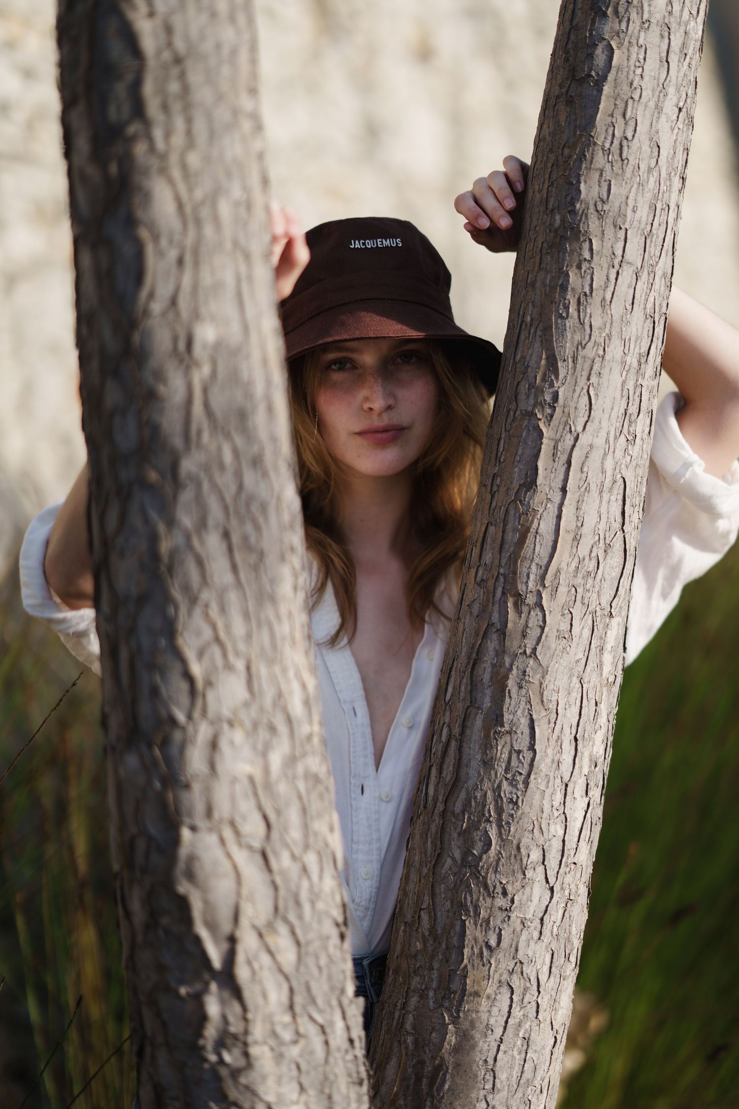 Young woman in a brown bucket hat and white top poses between two tree trunks in a natural outdoor setting.