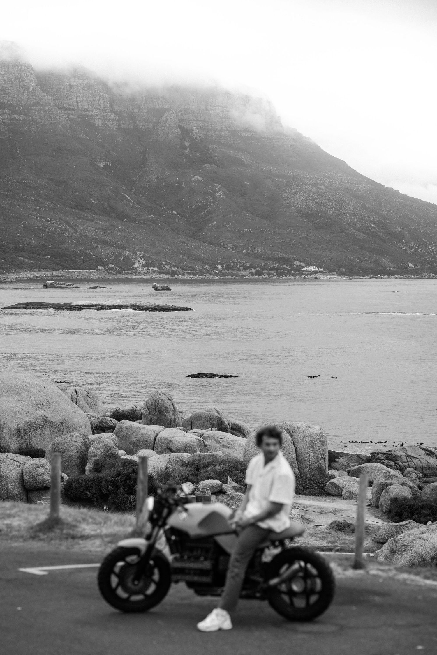 Black and white photo of a man leaning on a motorcycle by a rocky coastline with a misty mountain backdrop.
