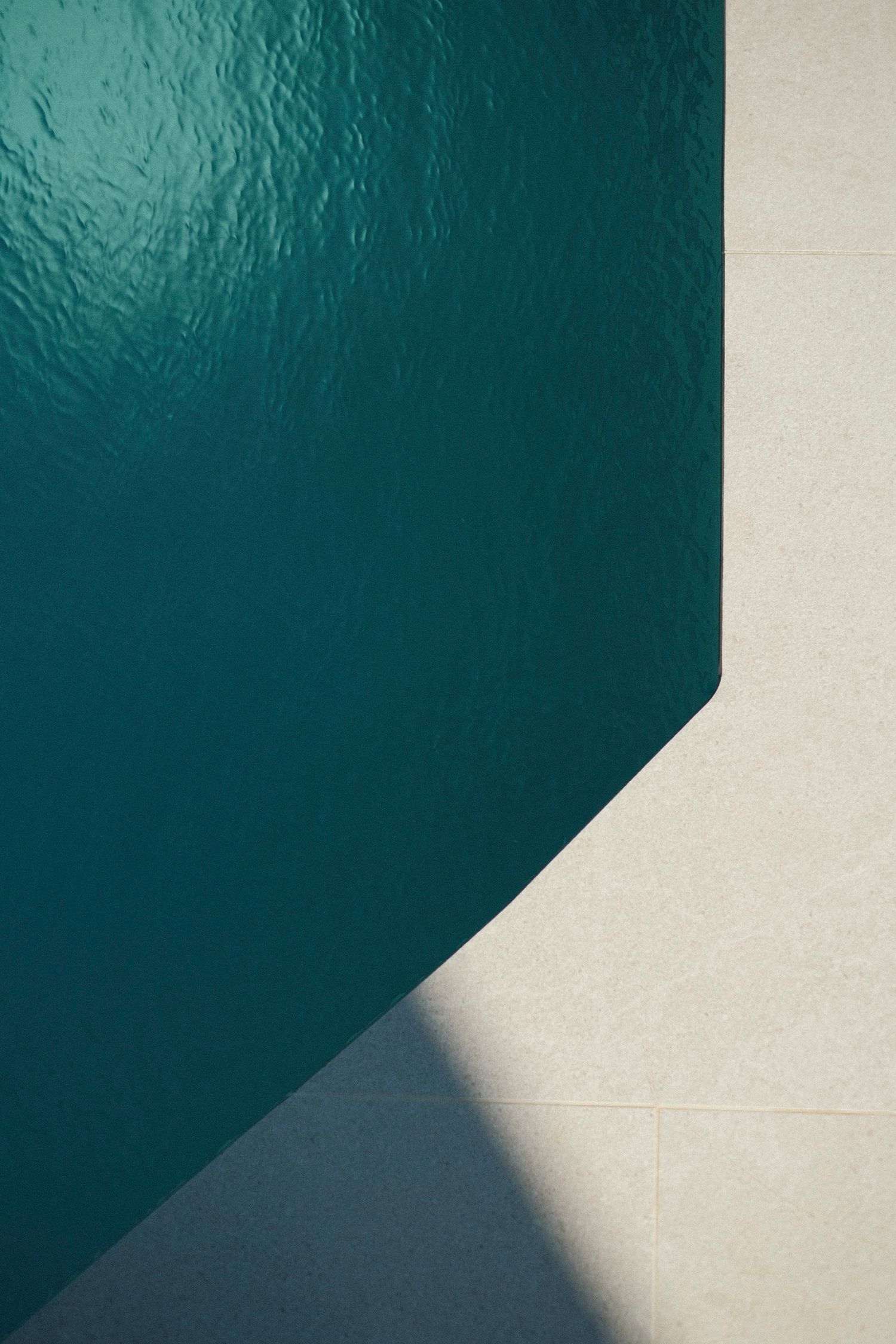 Aerial view of a teal swimming pool corner with beige stone tiles and geometric shadow patterns.