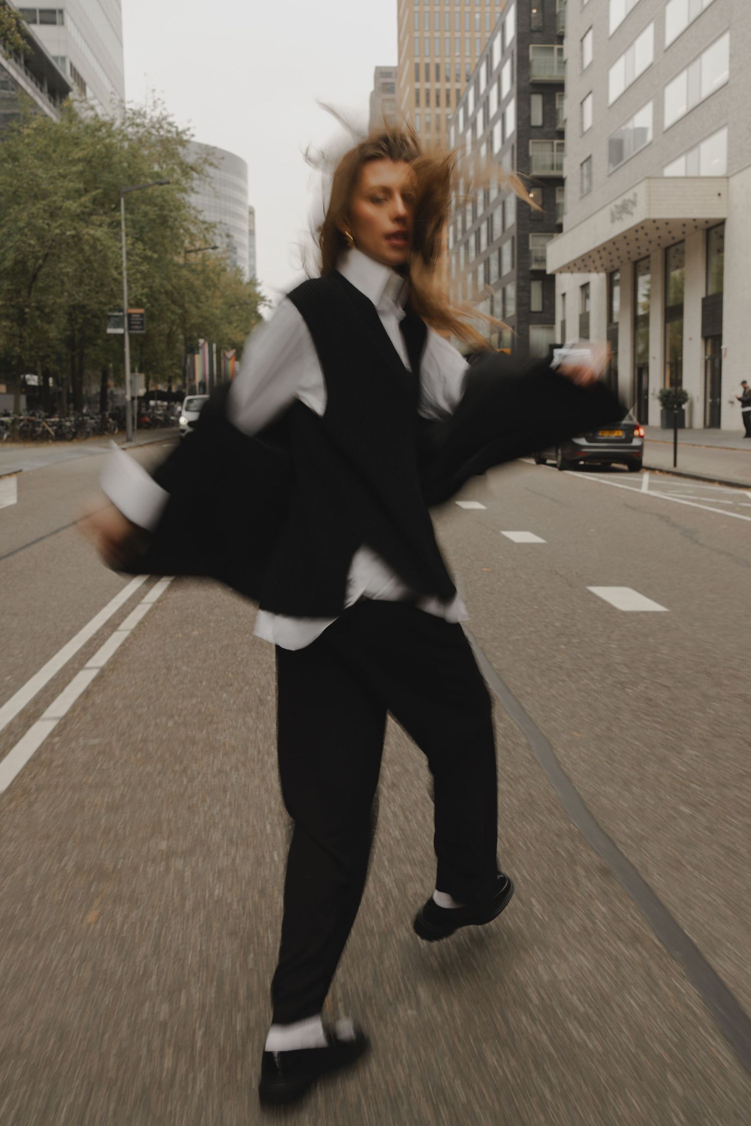 A woman in black trousers and vest dances dynamically on a city street, her hair and jacket blurred with motion.