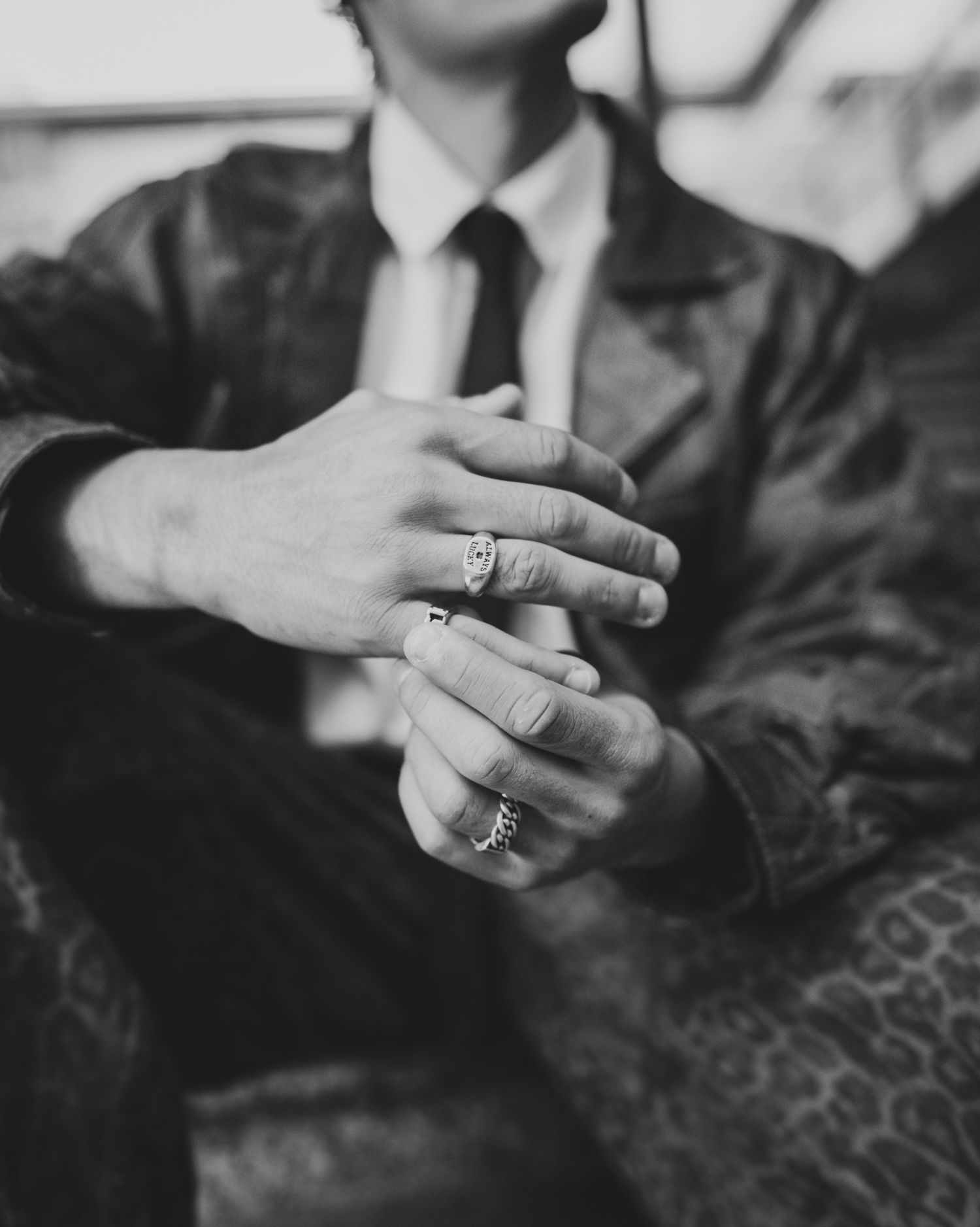 Black and white photo of a person in leather jacket and tie, hands clasped together wearing decorative rings.