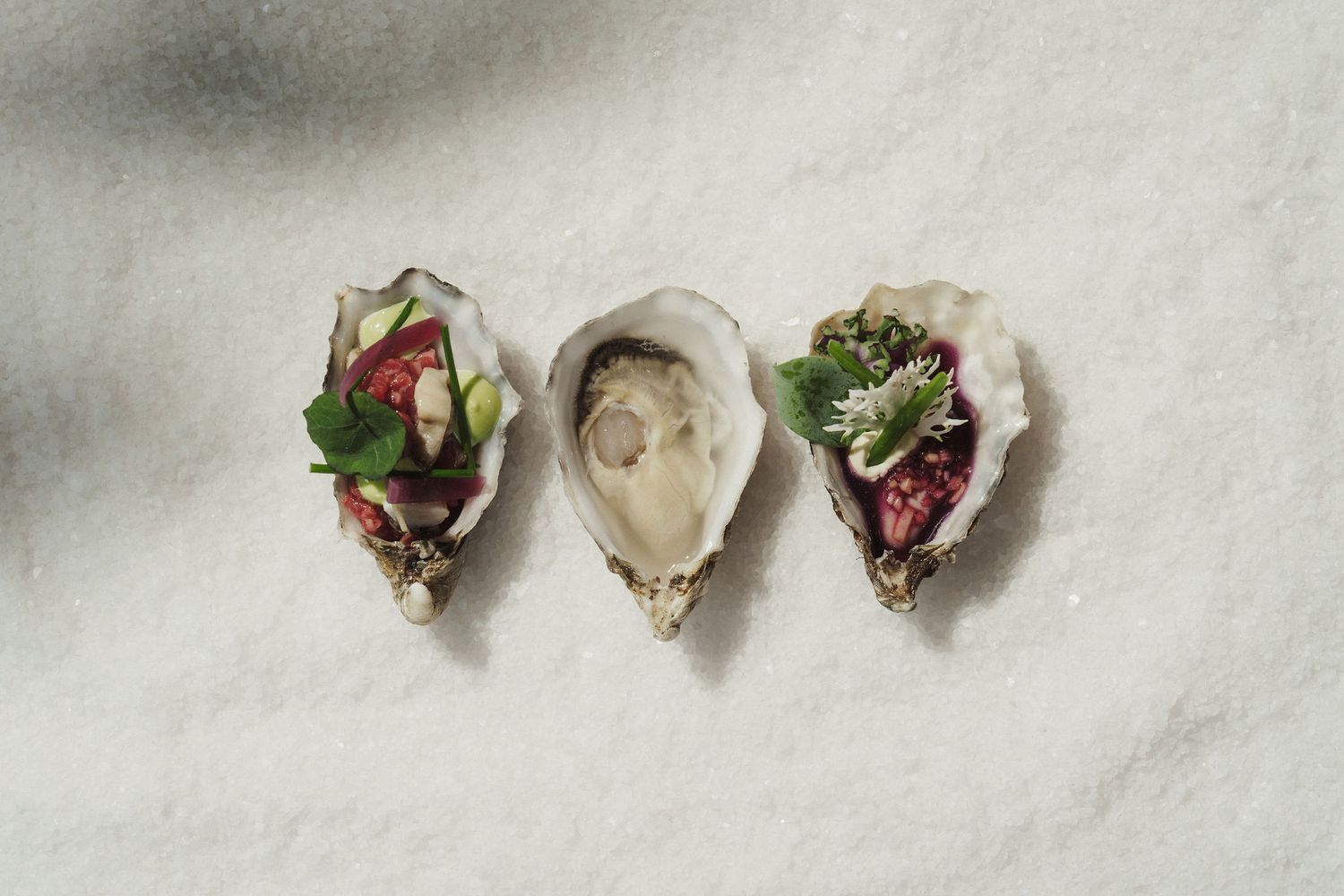 Three elegantly garnished oysters on the half shell with fresh herbs and flowers, displayed on a white surface.