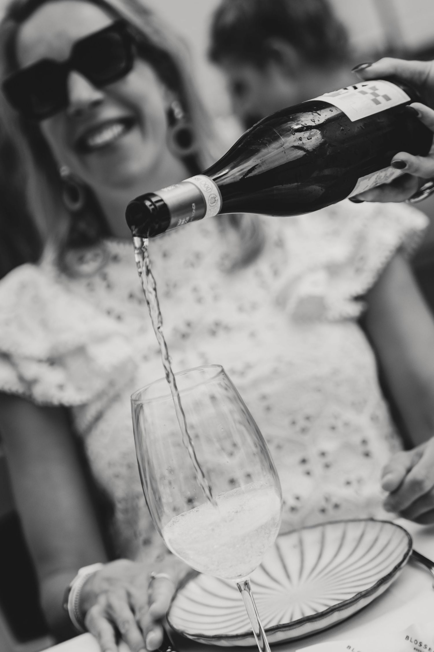 Black and white photo of wine being poured into a glass with a smiling woman in sunglasses in the background.