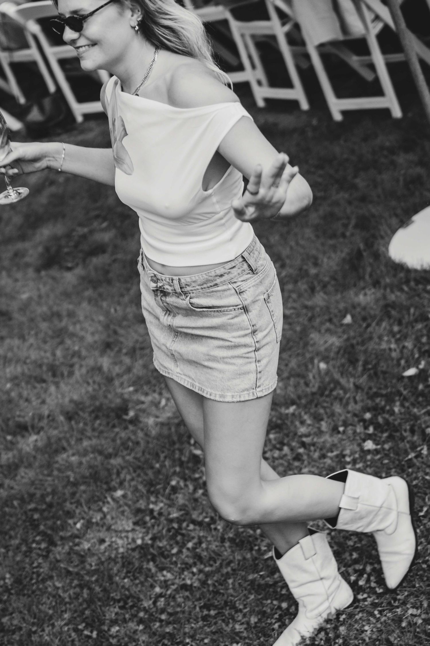 Black and white photo of a smiling woman in denim skirt and cowboy boots dancing outdoors at a garden party.