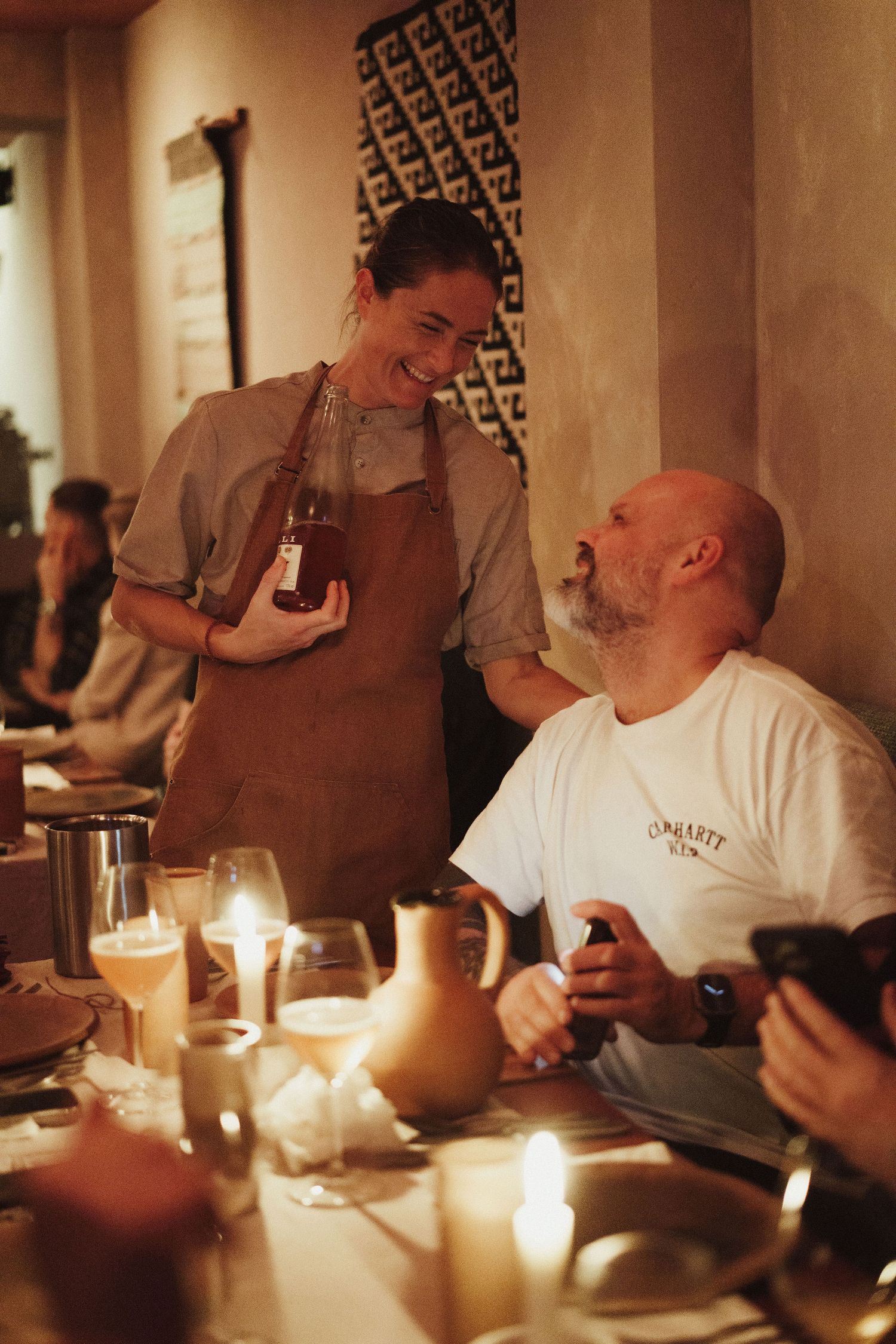 Server in brown apron smiles while holding a bottle, chatting with a bald bearded man at a candlelit restaurant table.