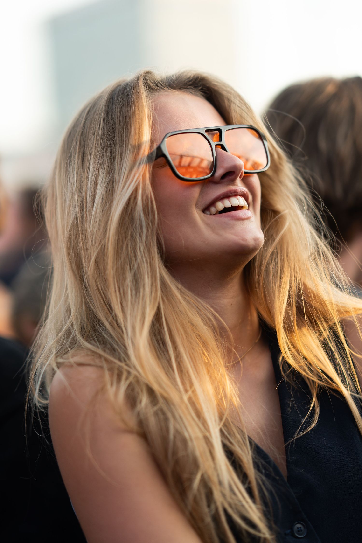 Young blonde woman laughing joyfully wearing orange mirrored sunglasses at an outdoor event in golden light.