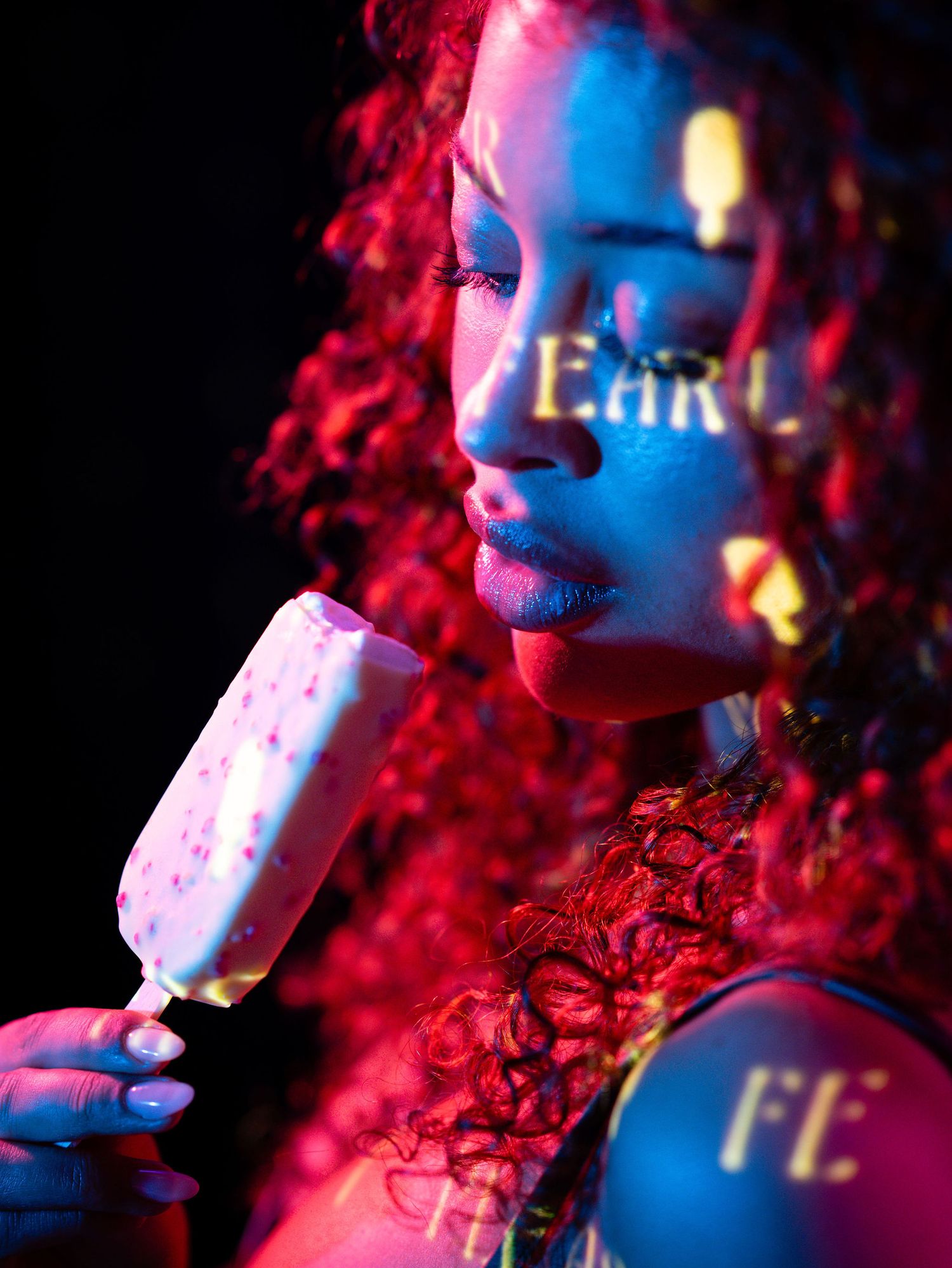 Woman with curly red hair licking an ice cream bar under neon pink and blue lights with projected text.
