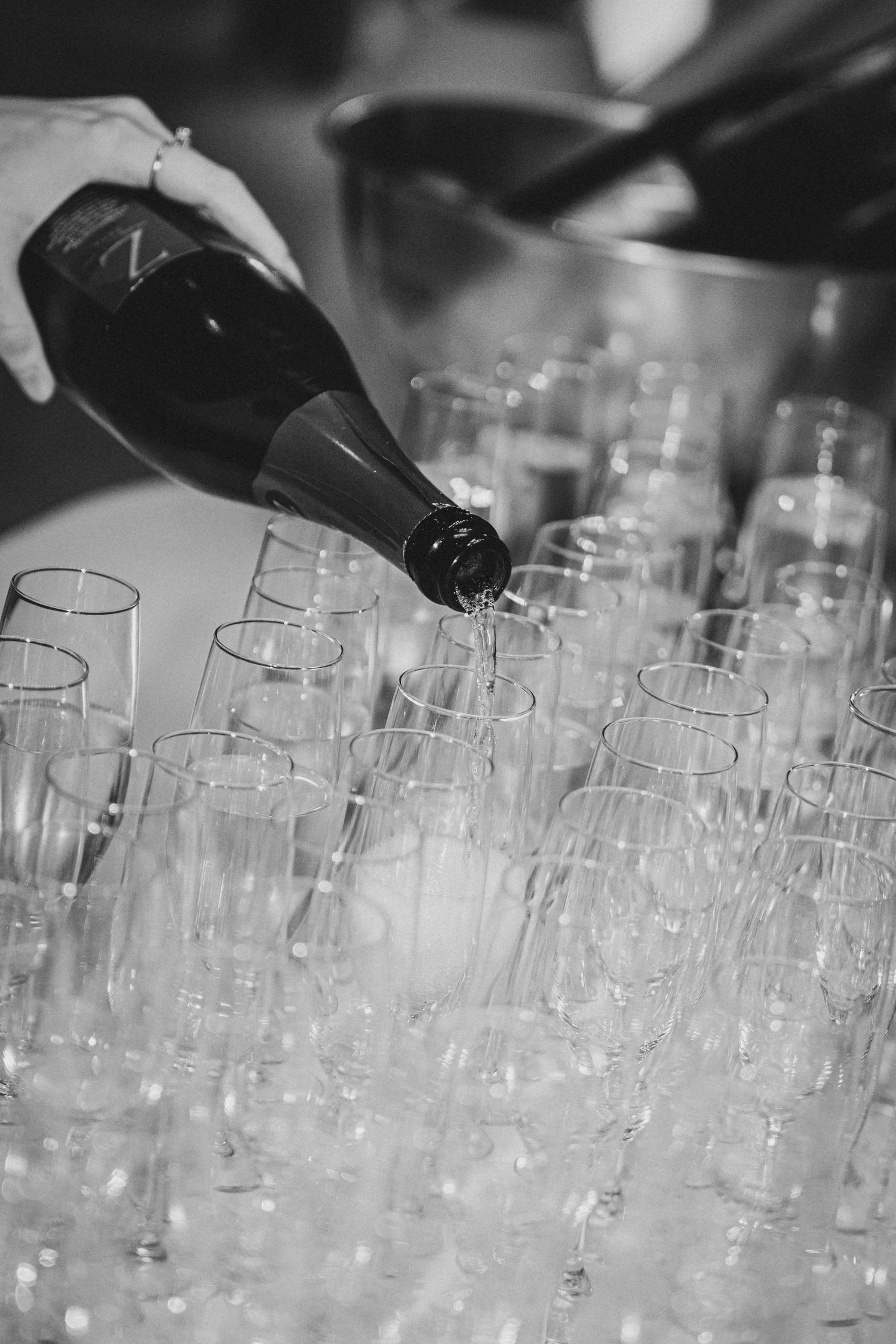 Black and white photo of champagne being poured into rows of elegant flute glasses at a formal event.
