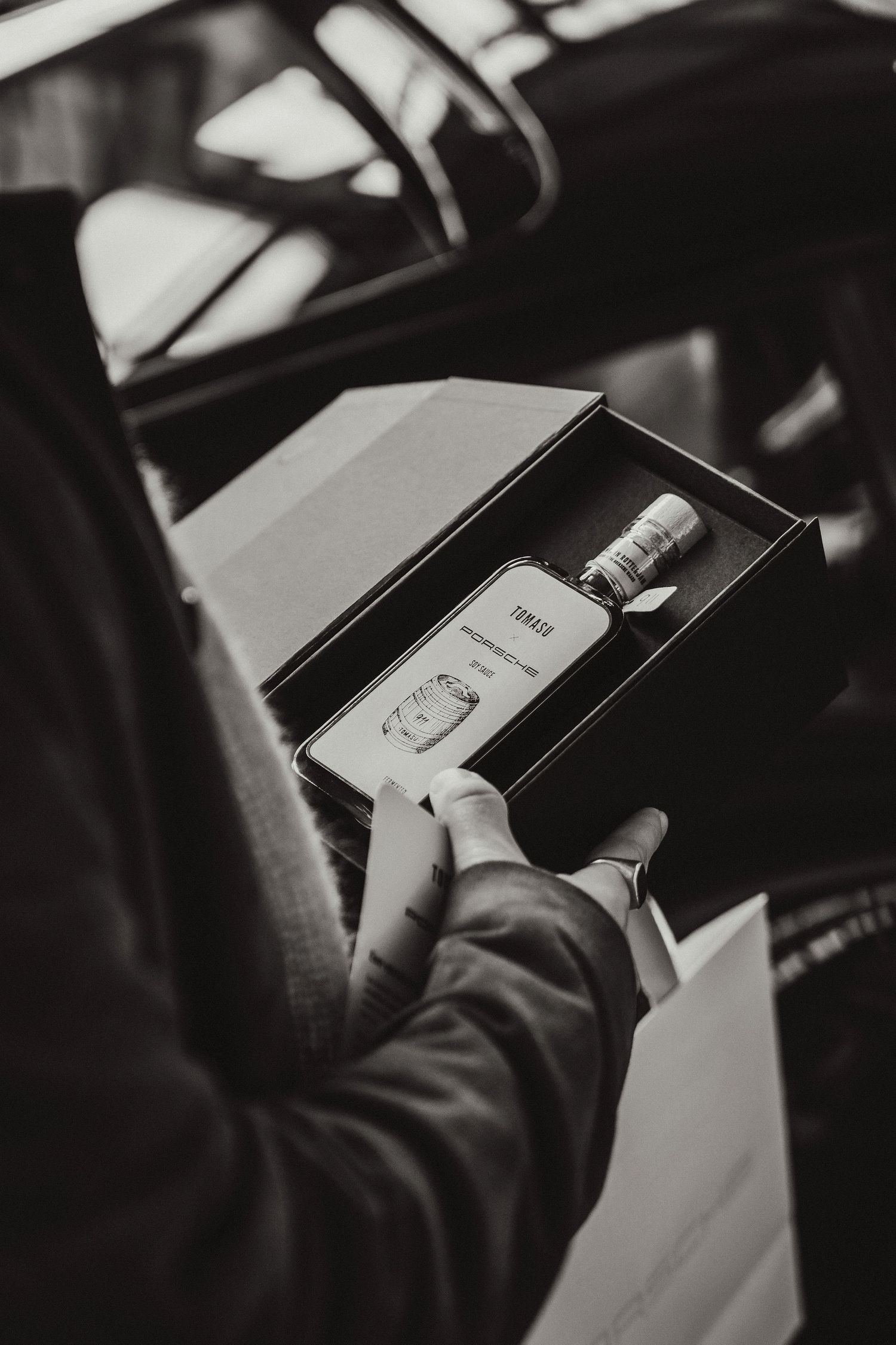 Black and white photo of hands holding an open gift box with a decorative bottle inside.