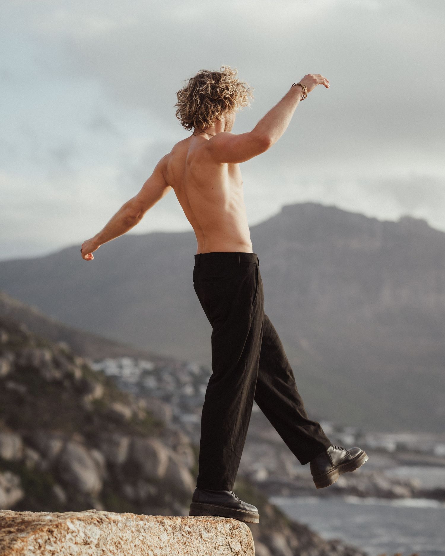 Person dancing on coastal cliff edge with mountains and ocean in background.