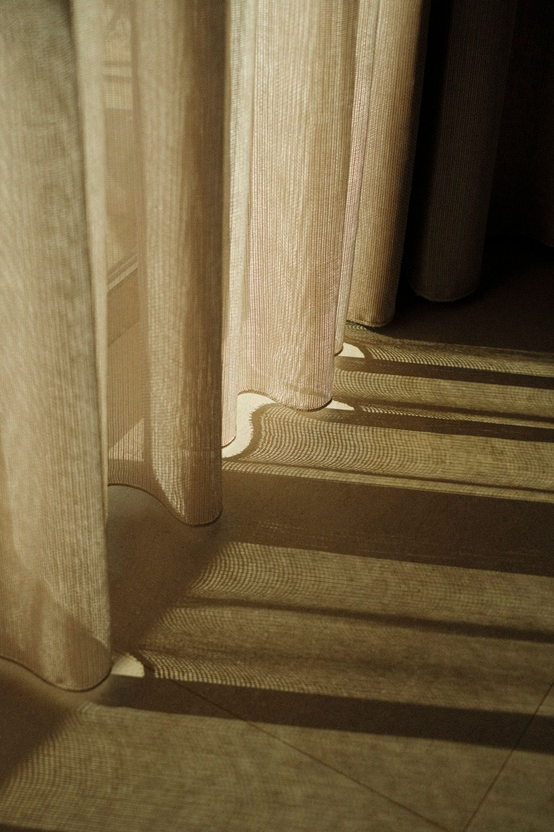 Sheer beige curtains casting soft striped shadows on a tiled floor in warm golden afternoon light.