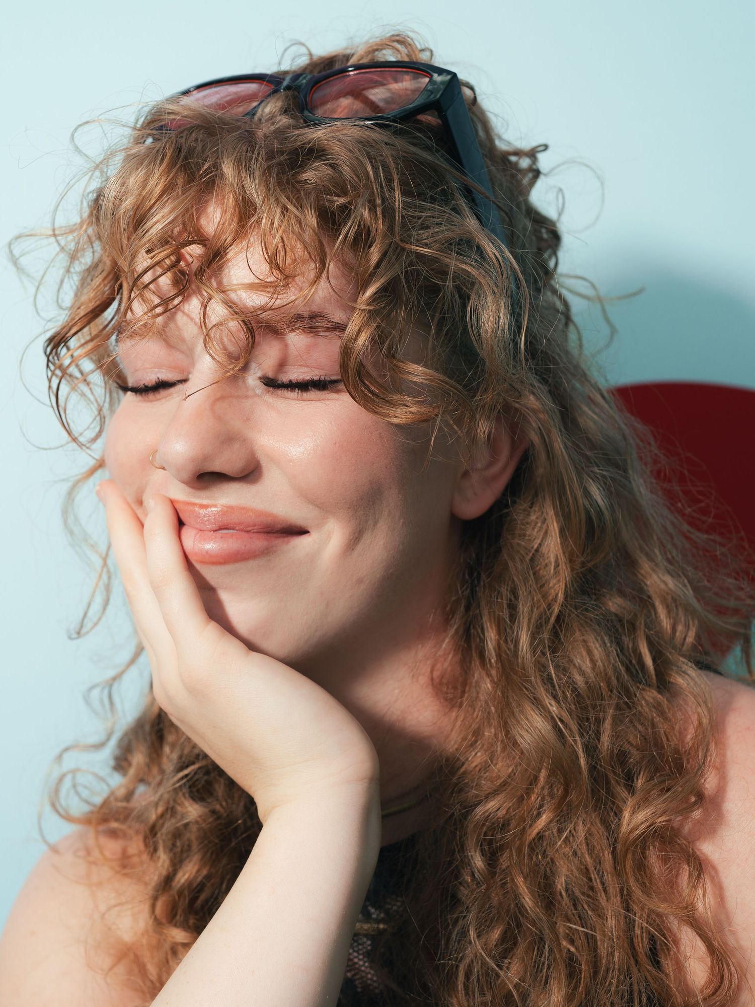Young woman with curly red hair and sunglasses on head, eyes closed, resting cheek on hand against blue background.
