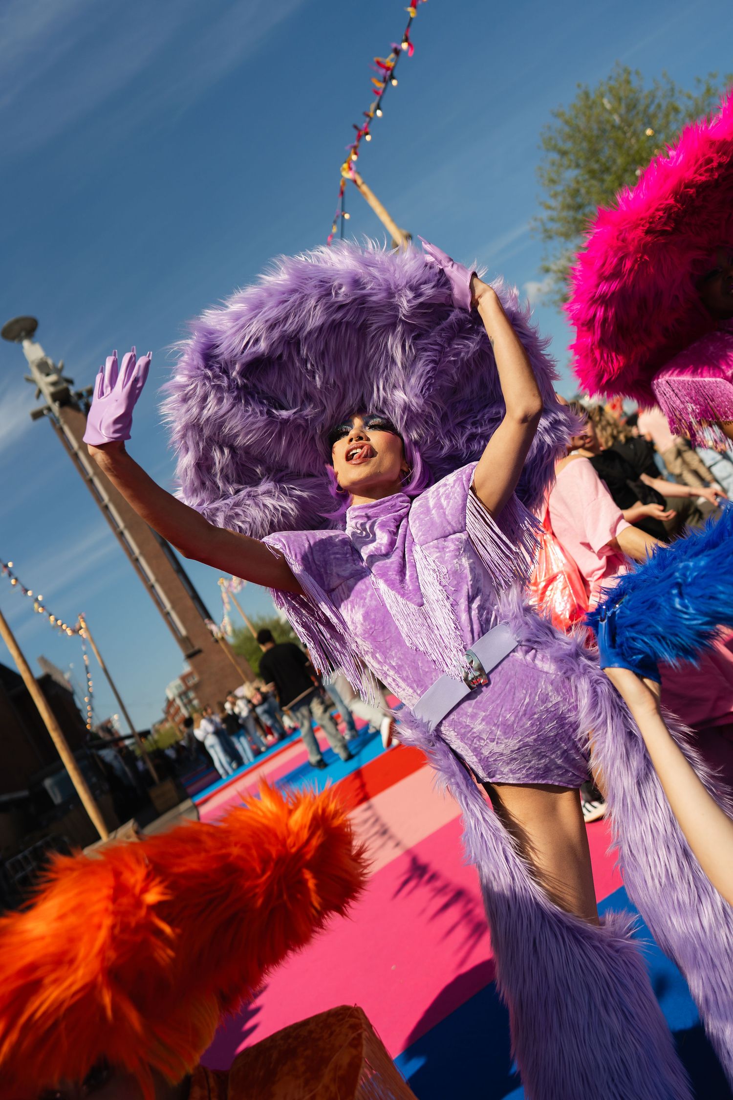 Performer in vibrant purple feathered costume dancing at a colorful outdoor carnival festival under blue sky.