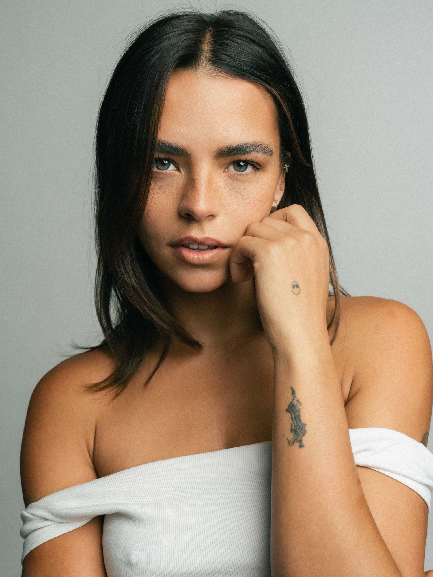 Young woman with freckles and dark hair in white off-shoulder top, hand raised to chin, wrist tattoo.