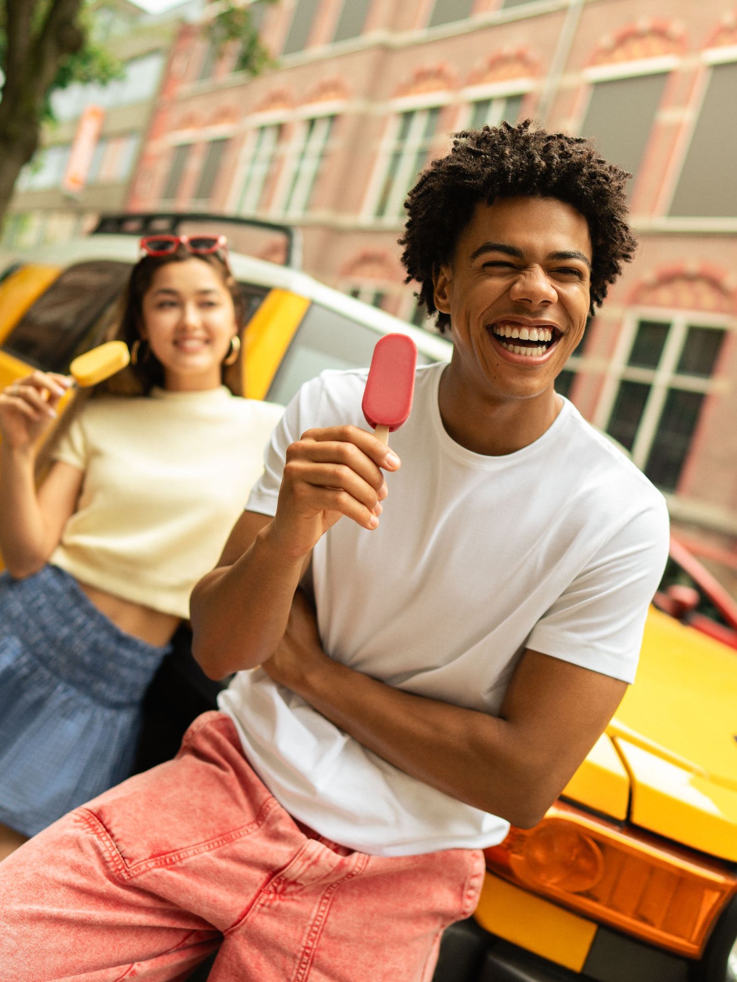Two teens laughing and eating colorful ice cream bars outdoors near a yellow car on a sunny day.
