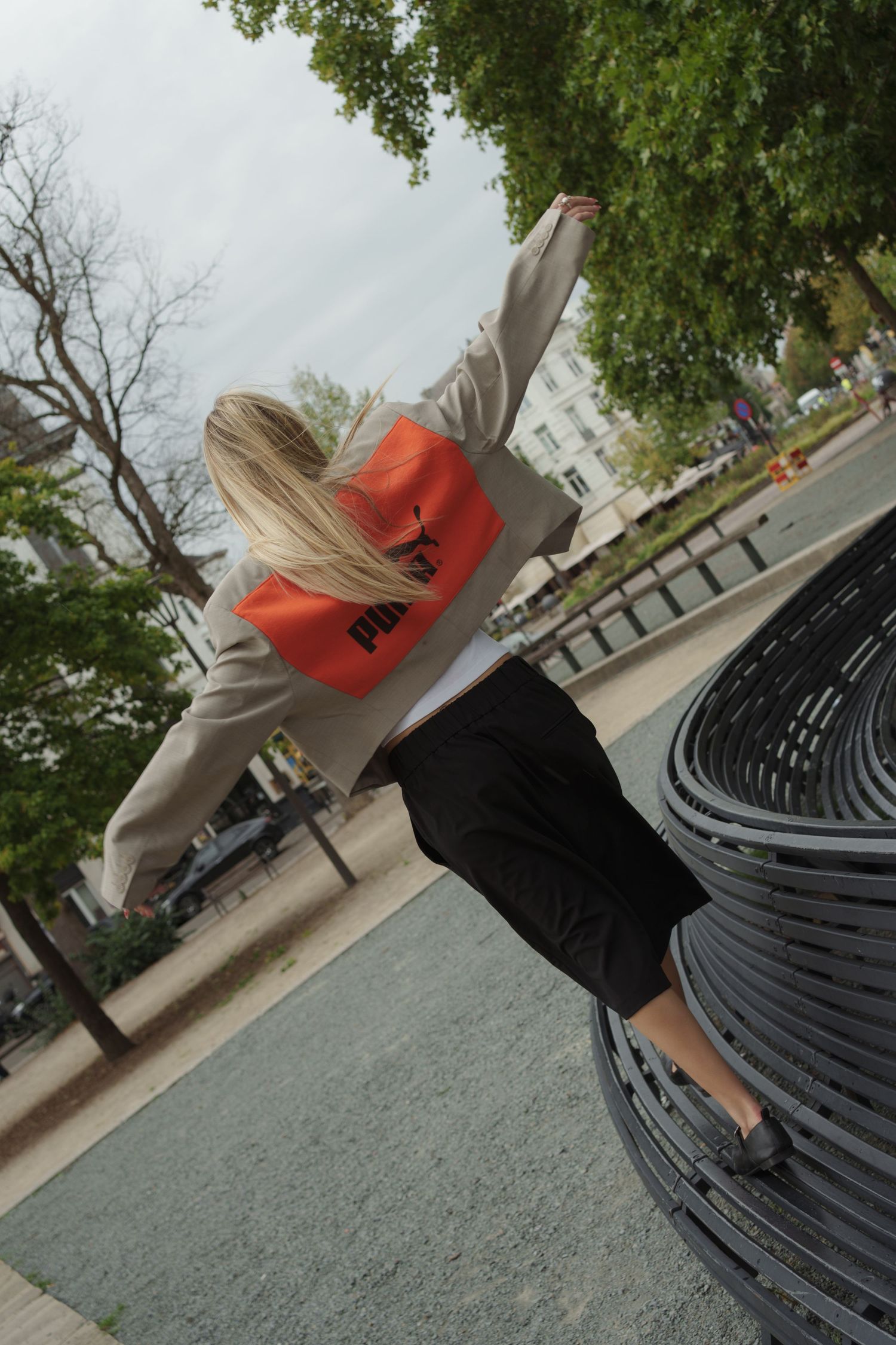 Blonde woman in beige blazer with orange Puma bag raising arm in urban park setting, rear view.
