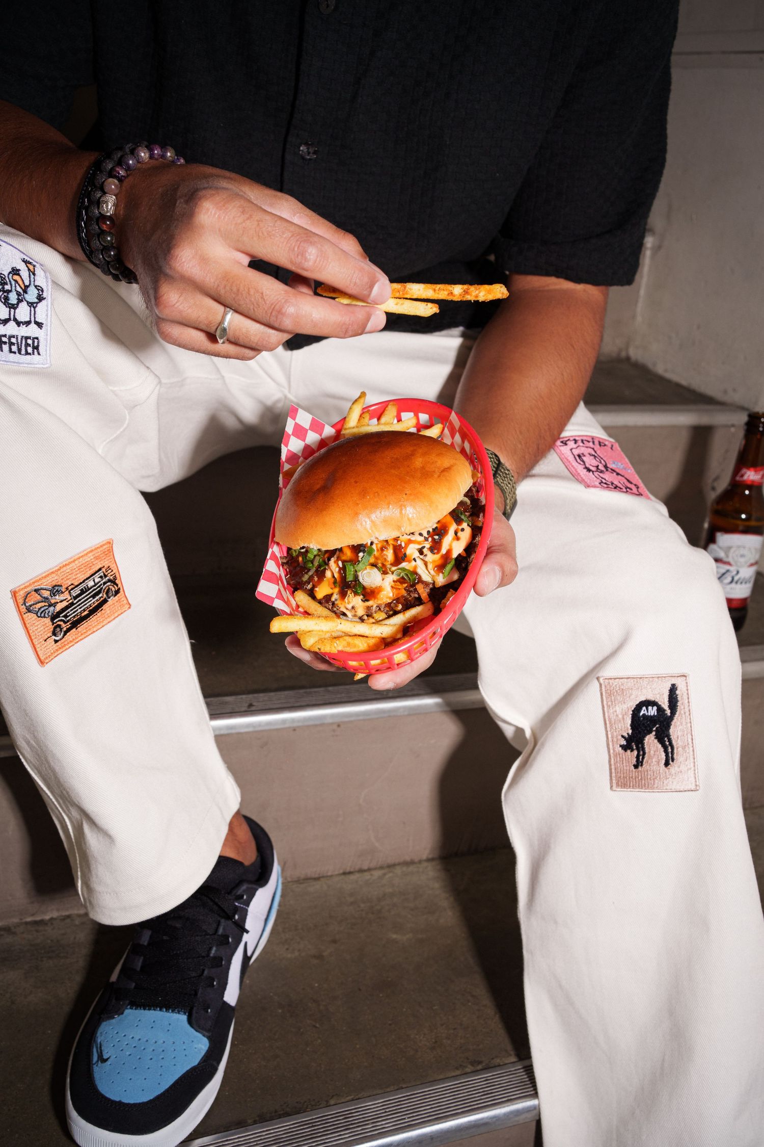 Person holding a loaded burger and fries in a red checkered basket, wearing white pants with patches and Jordan sneakers.