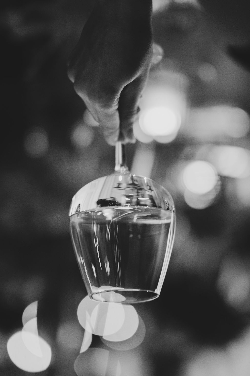 Black and white photo of a hand holding an inverted wine glass with bokeh lights in the background.