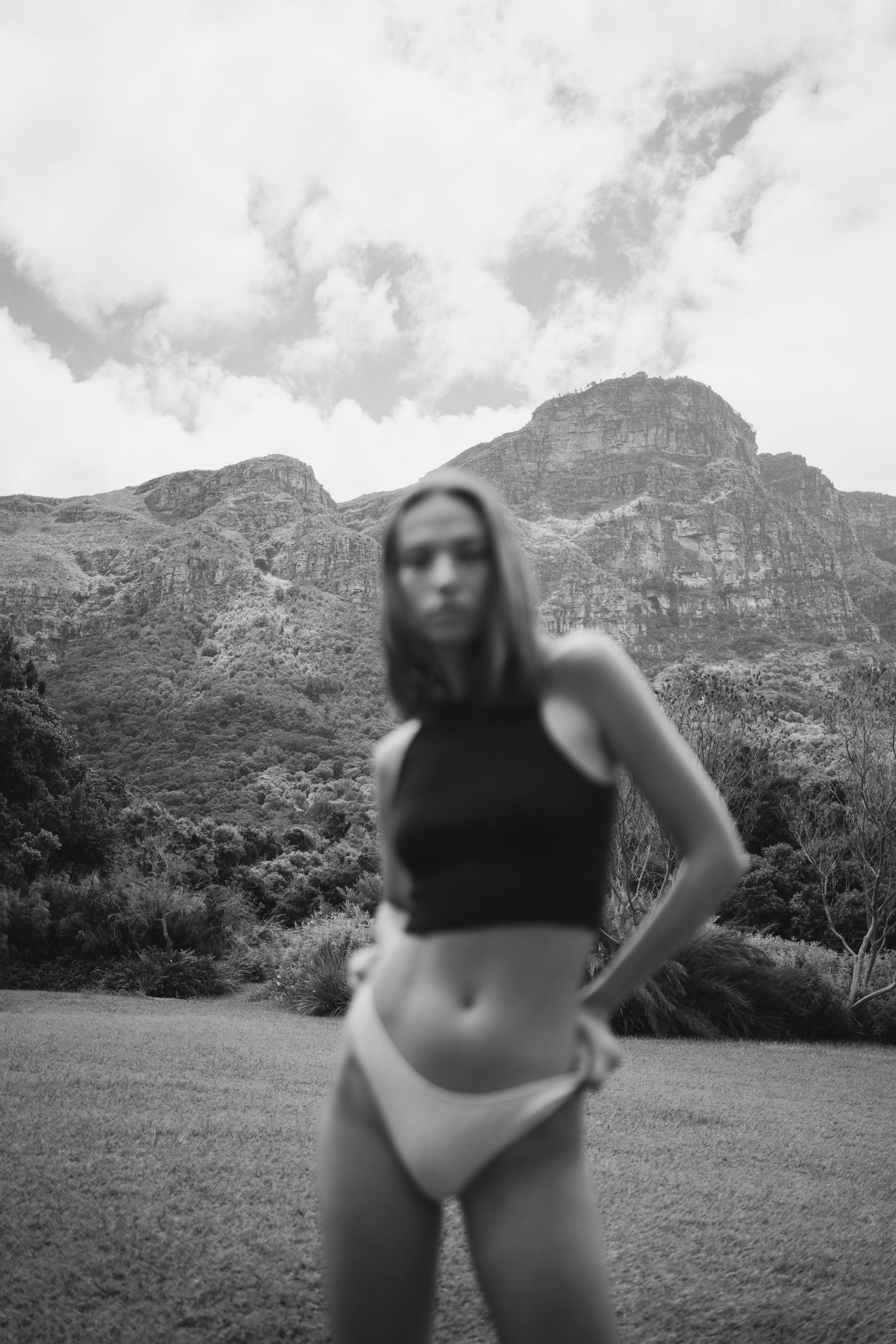 Black and white photo of a woman in crop top and bikini bottoms posing outdoors with mountains behind her.