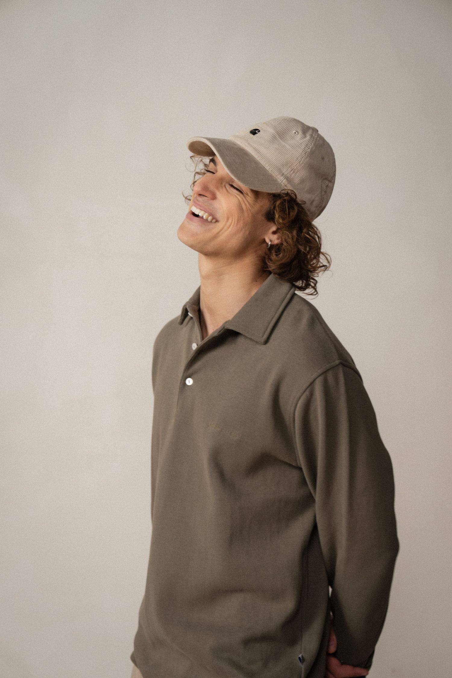 A person wearing a brown sweatshirt and baseball cap laughs joyfully while looking upward in a studio setting.