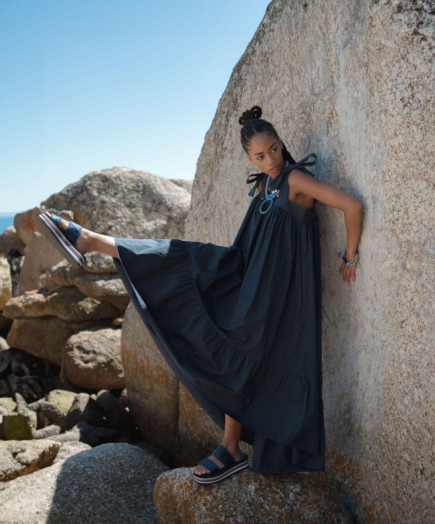 Woman in black maxi dress and platform sandals posing against granite rocks at a sunny coastal location.
