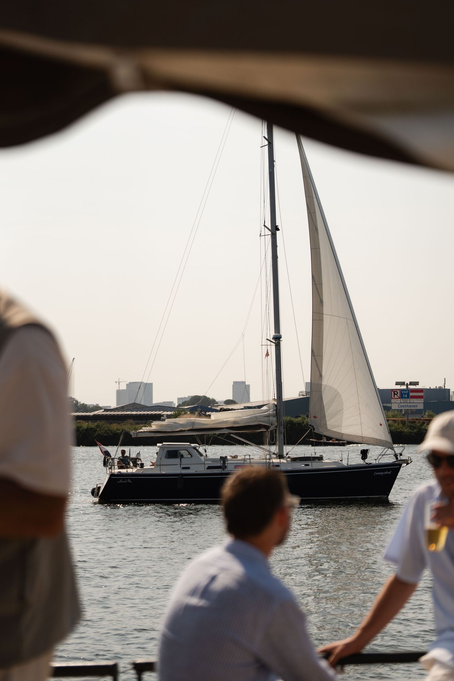 A sailboat glides along a calm harbor waterway as people gather on a nearby dock on a sunny day.