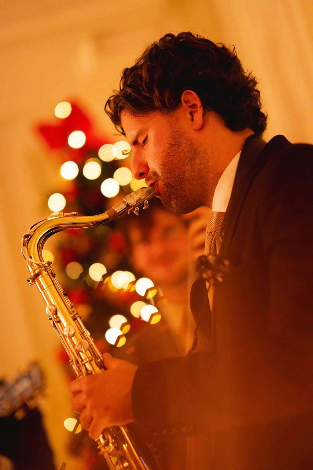 Man playing saxophone at a Christmas party with festive bokeh lights and decorated tree in background.