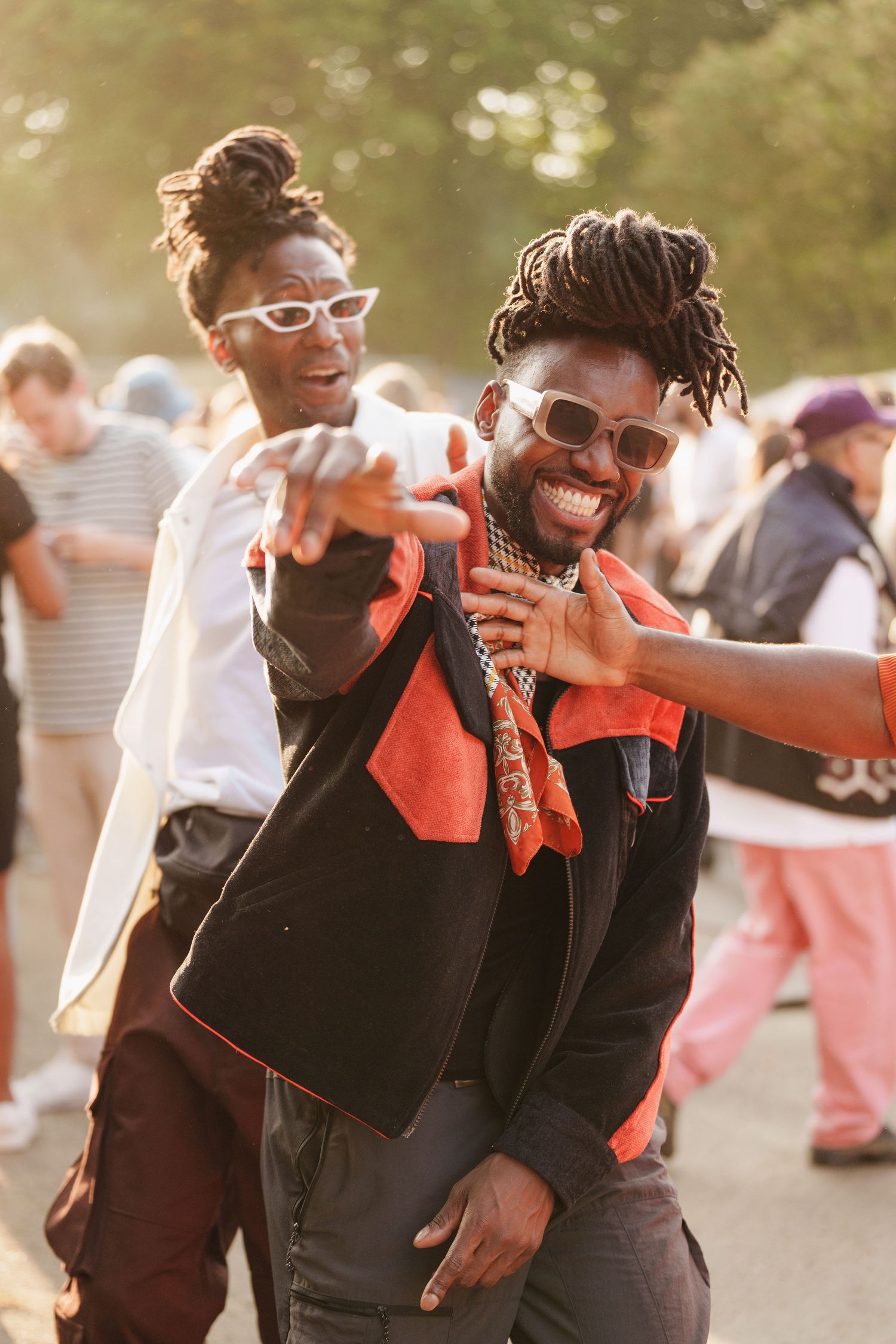 Two stylish Black men dancing and pointing at a sunny outdoor festival, wearing sunglasses and trendy outfits.