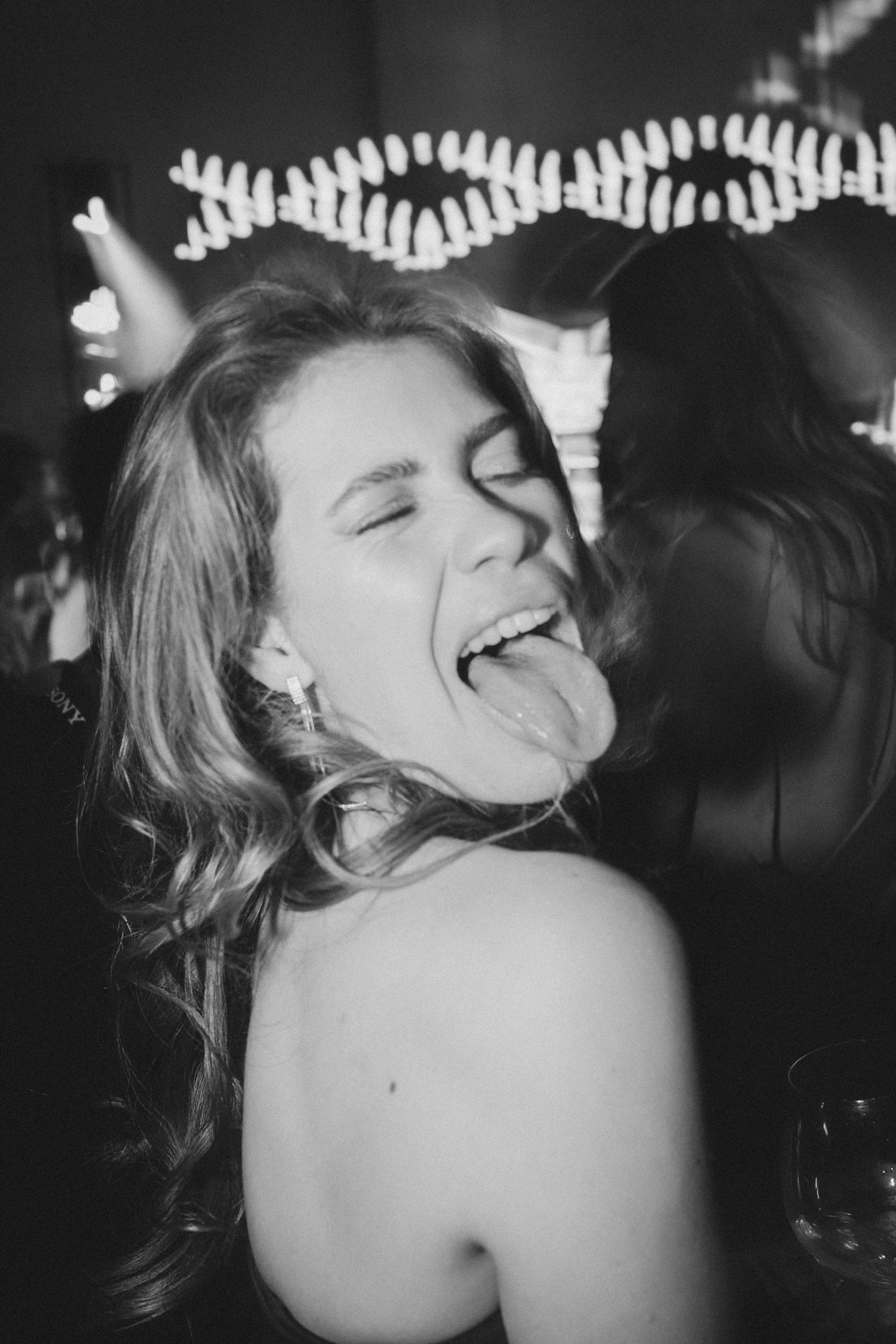 Black and white photo of a young woman laughing with tongue out at a lively party event.